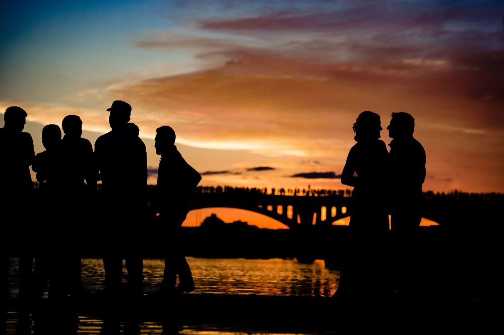 Silhouettes of wedding guests gathered at sunset with a bridge in the background.