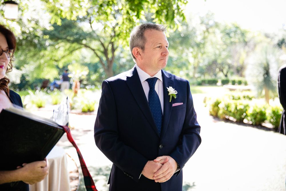 A groom in a navy suit stands outdoors during a sunny wedding ceremony.