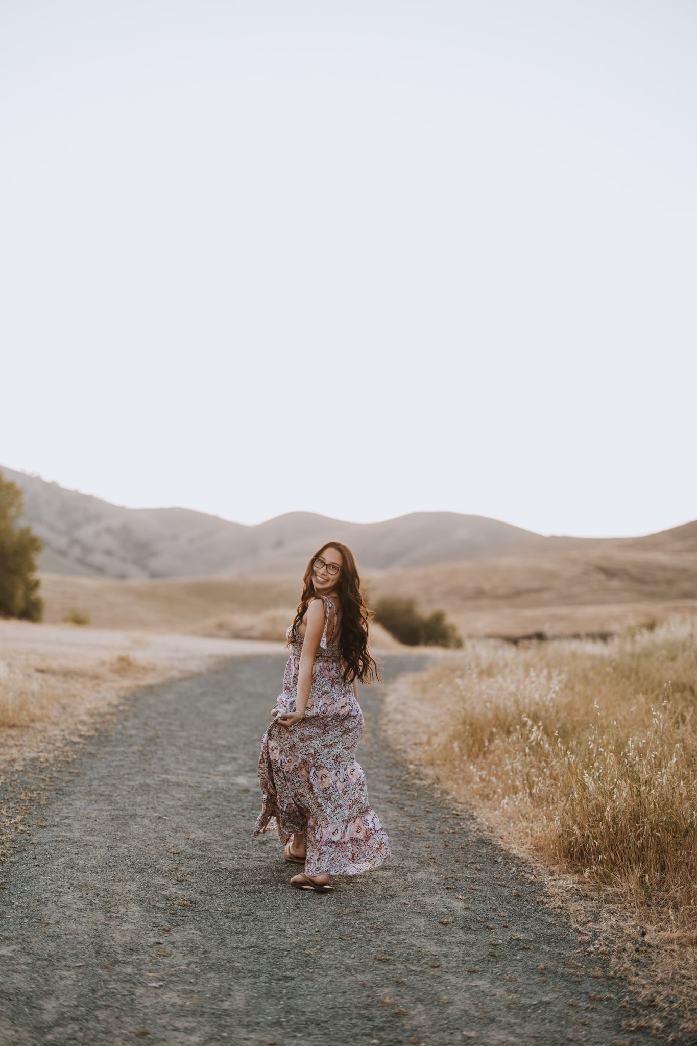 Woman in floral maxi dress walks along a dirt path with mountains in the background at sunset.