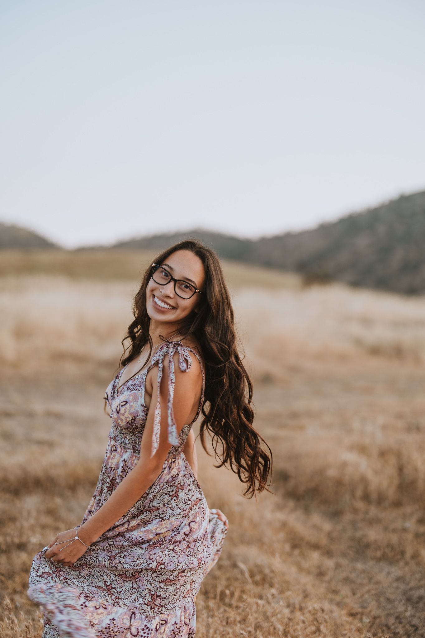 Woman in floral dress twirling joyfully in a sun-dried field with mountains behind.