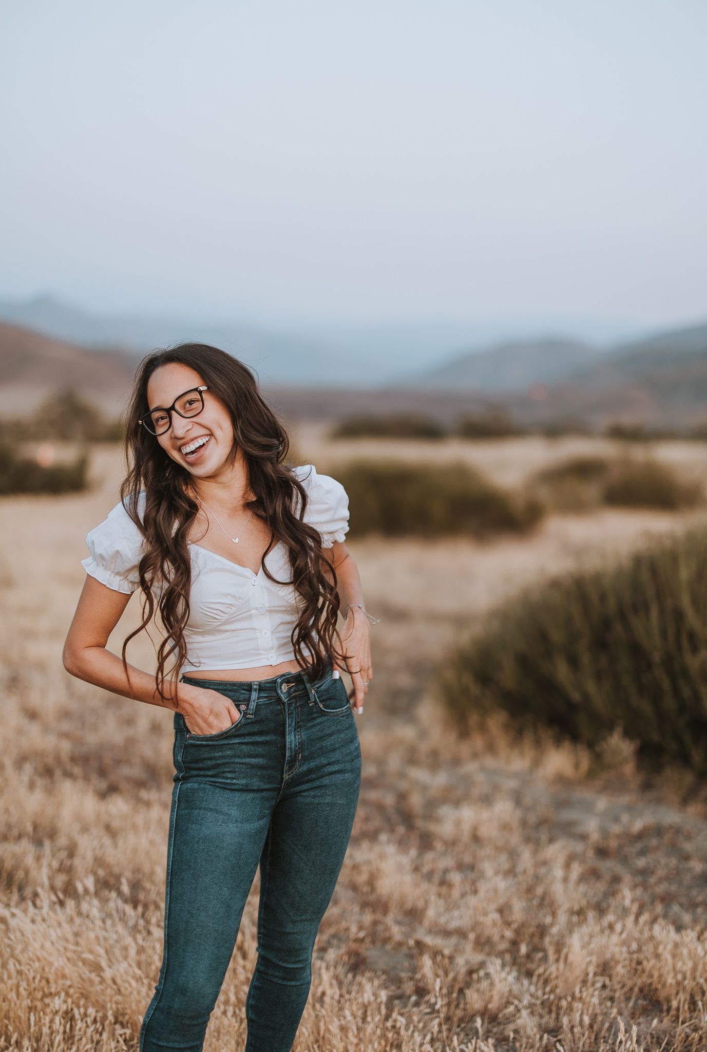 Young woman in casual attire poses playfully against golden hills and mountains backdrop.