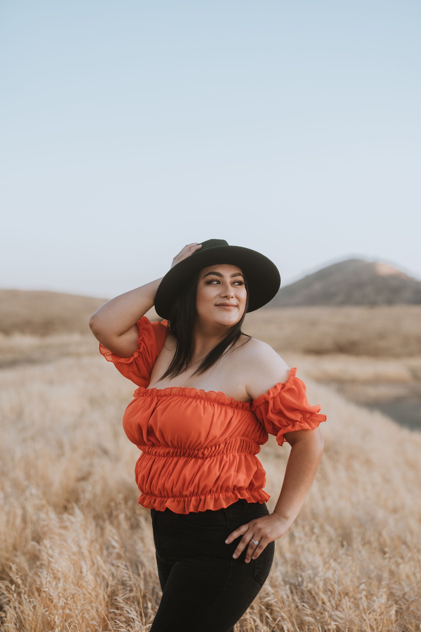 Person wearing orange off-shoulder top and black hat poses in dry grass field.