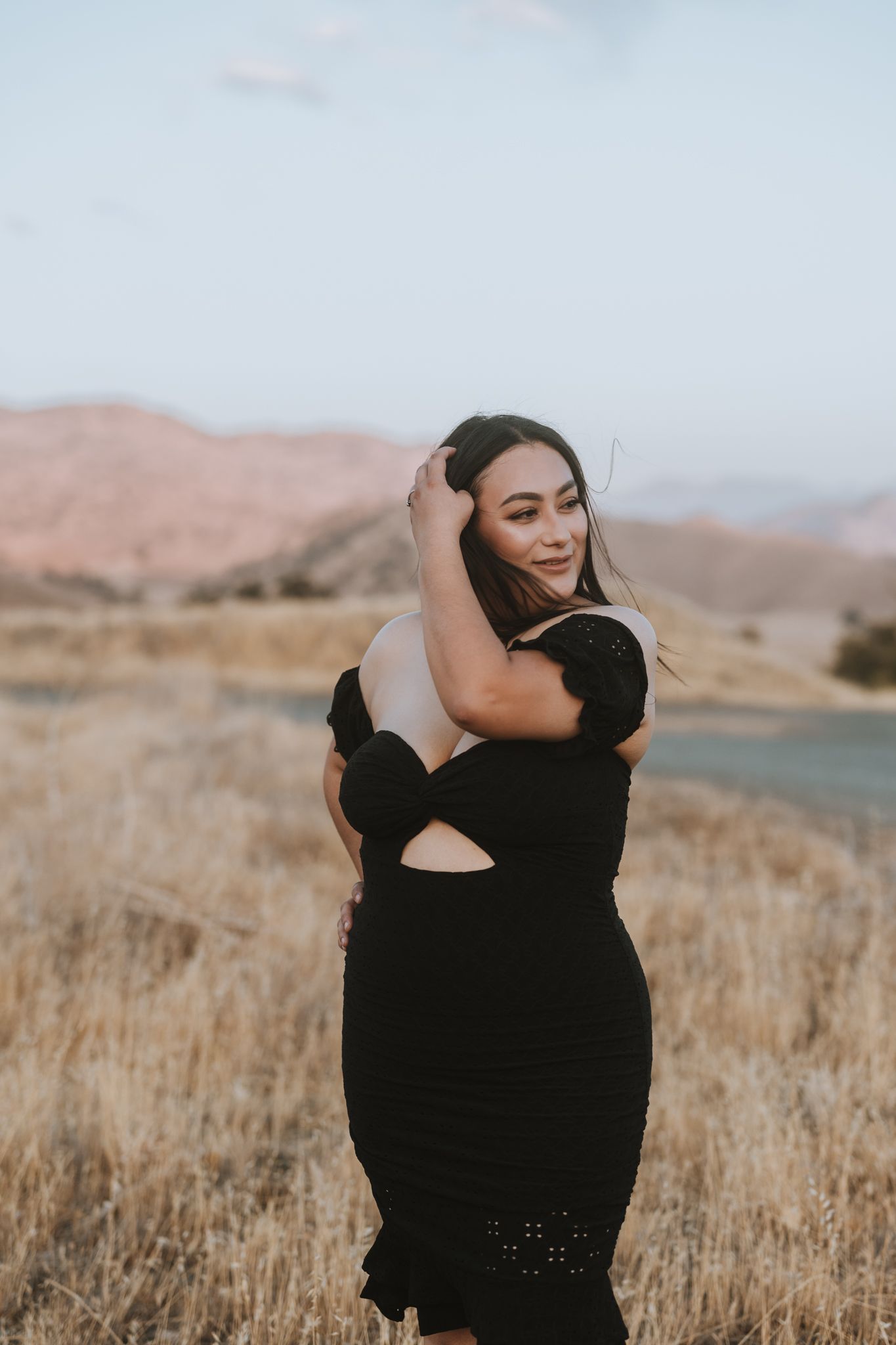 Person in black dress poses gracefully in field with mountains in background.