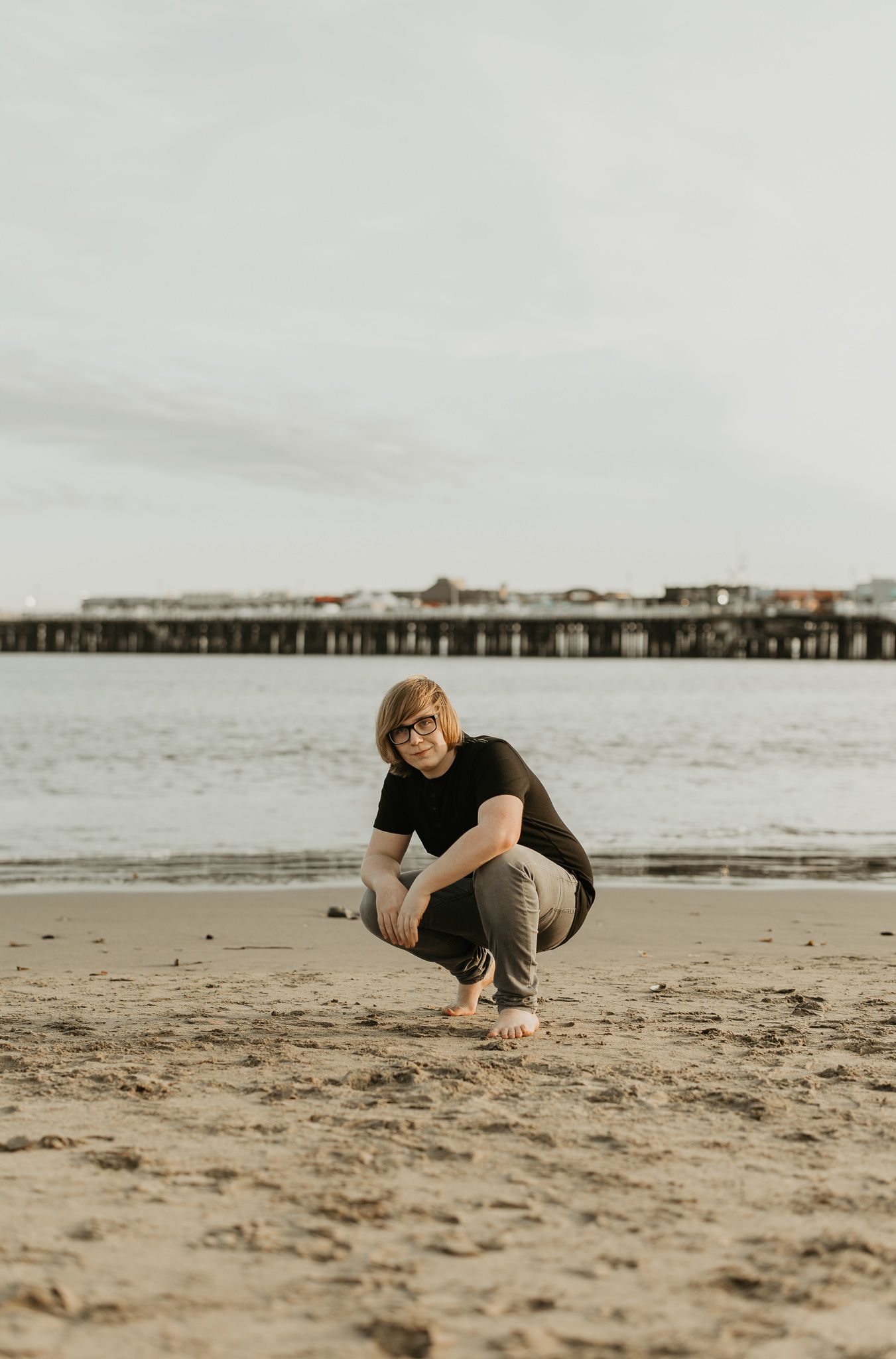 Person crouching on beach with pier visible in distance.