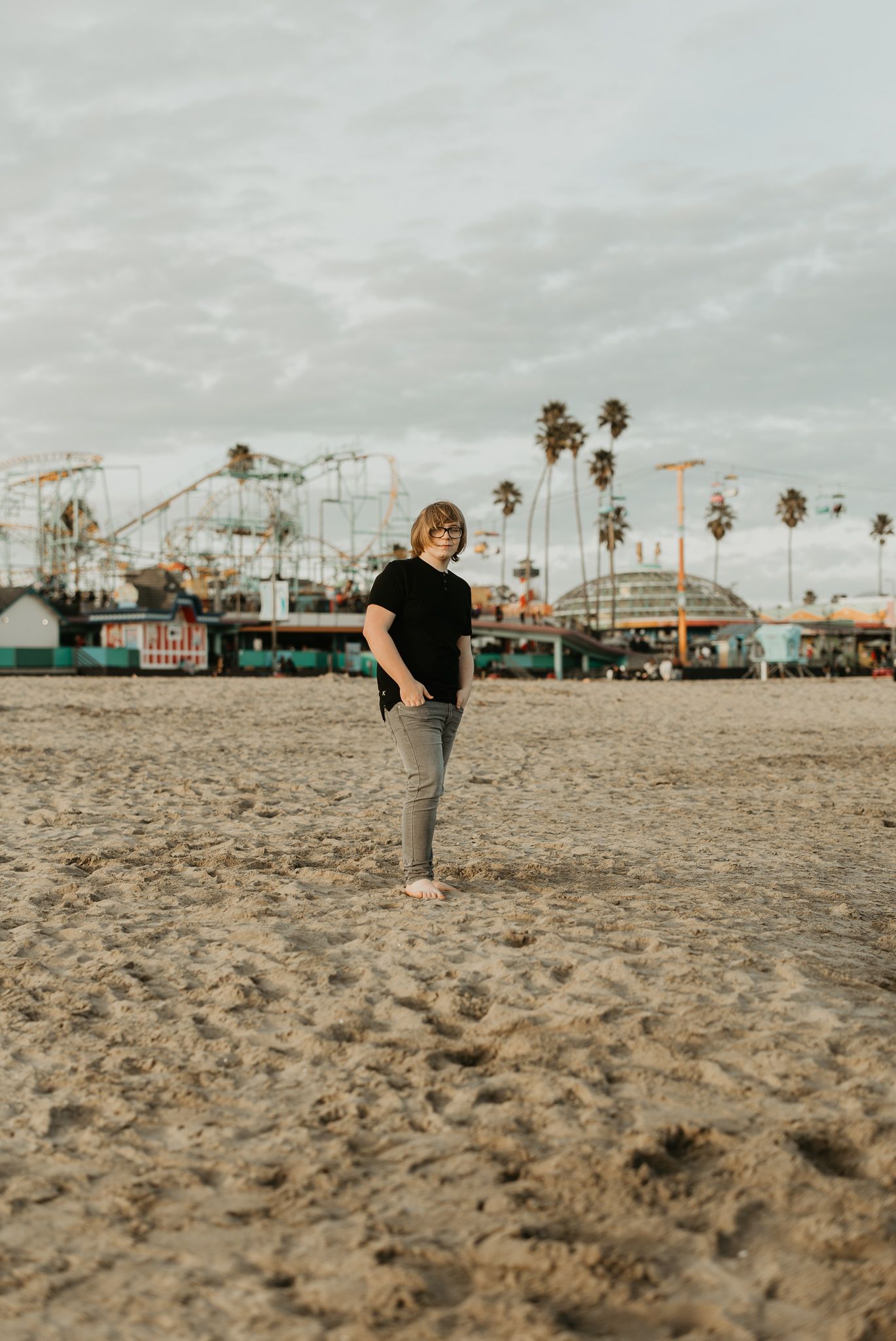 Person standing on sandy beach with amusement park rides and palm trees in background.