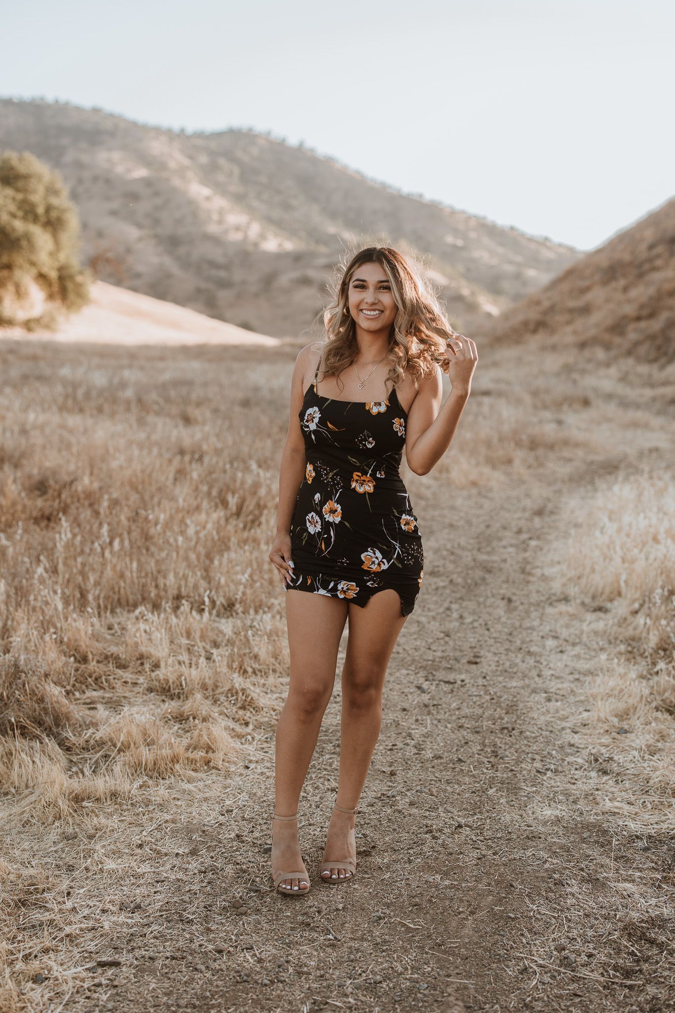 Woman in floral romper poses on dirt path with mountains in background during golden hour.