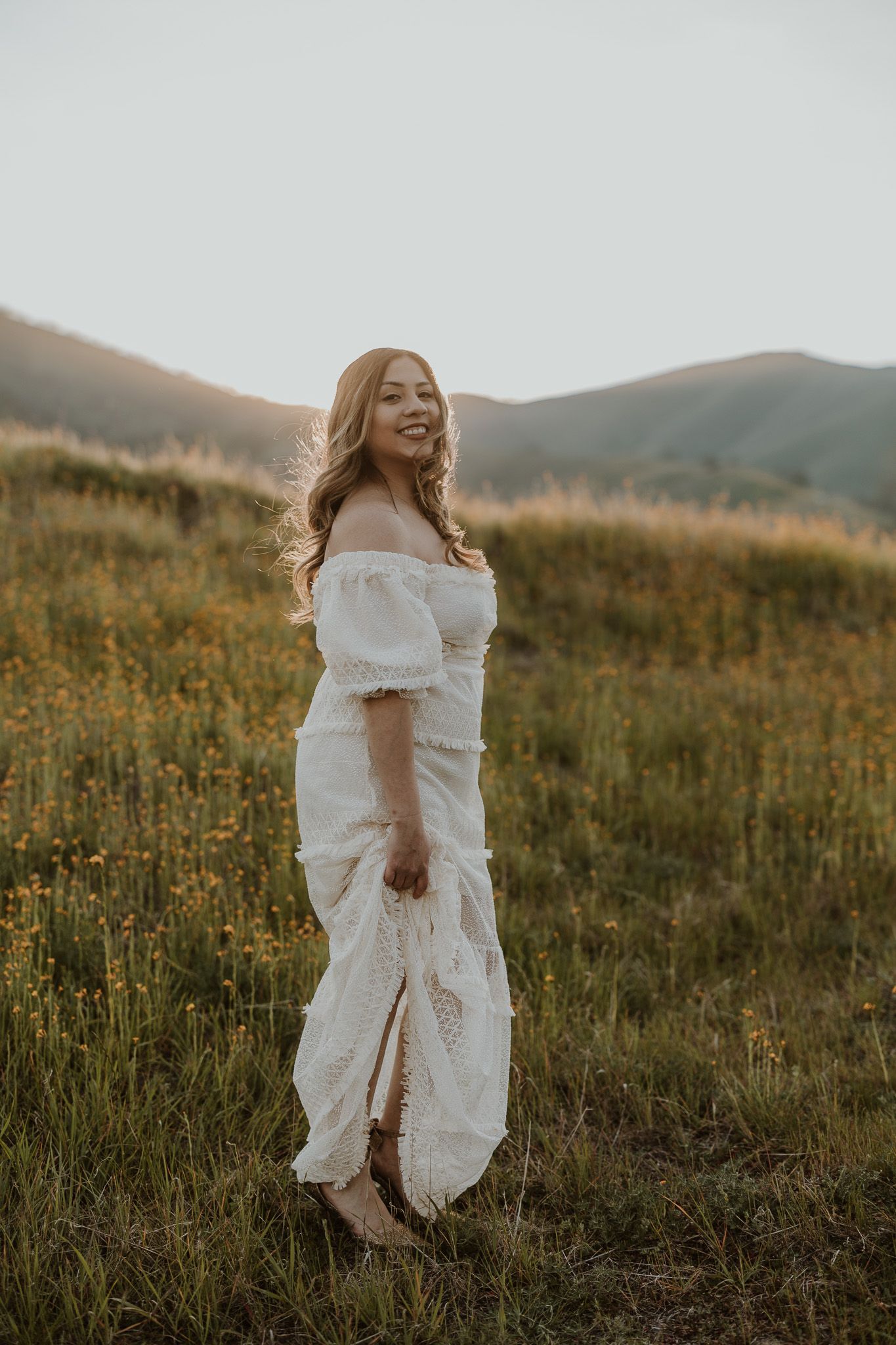 Woman in flowing white dress poses gracefully in mountain meadow at golden hour.