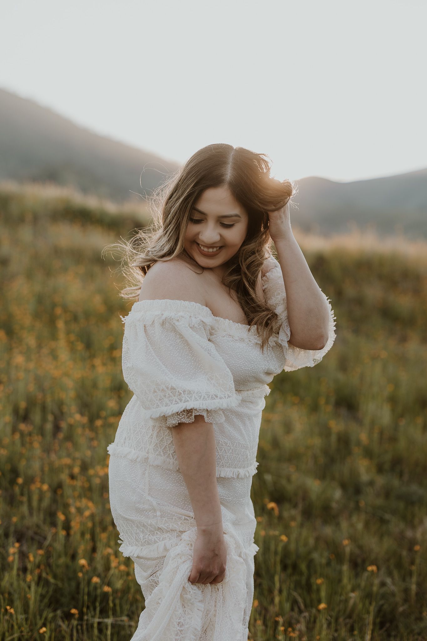 Woman in white off-shoulder dress smiling joyfully in a wildflower meadow at sunset.
