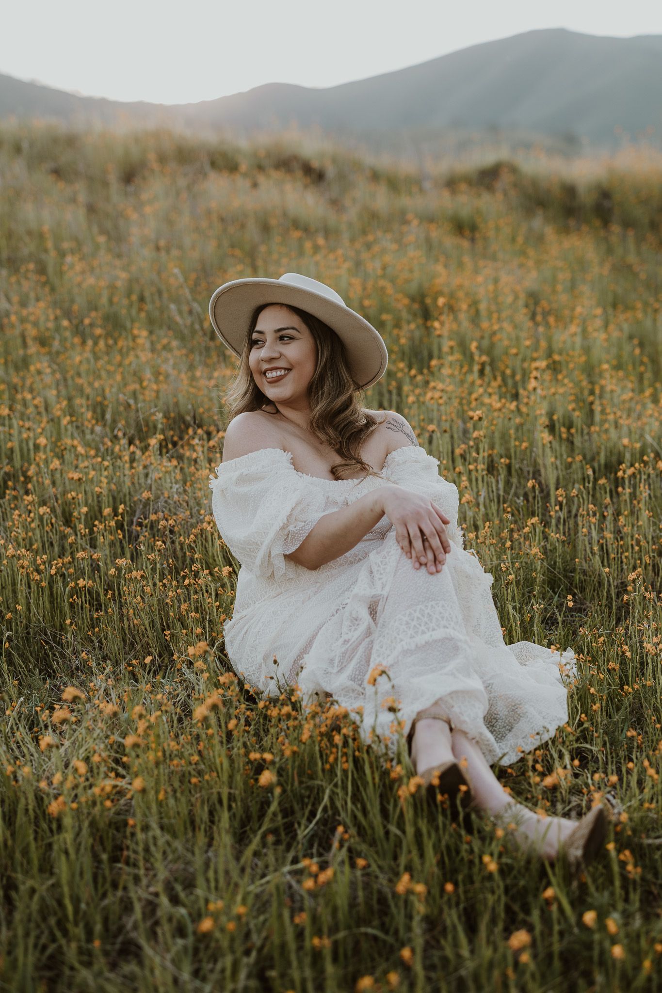Woman in white dress and sun hat sitting in a blooming wildflower field with mountains behind.