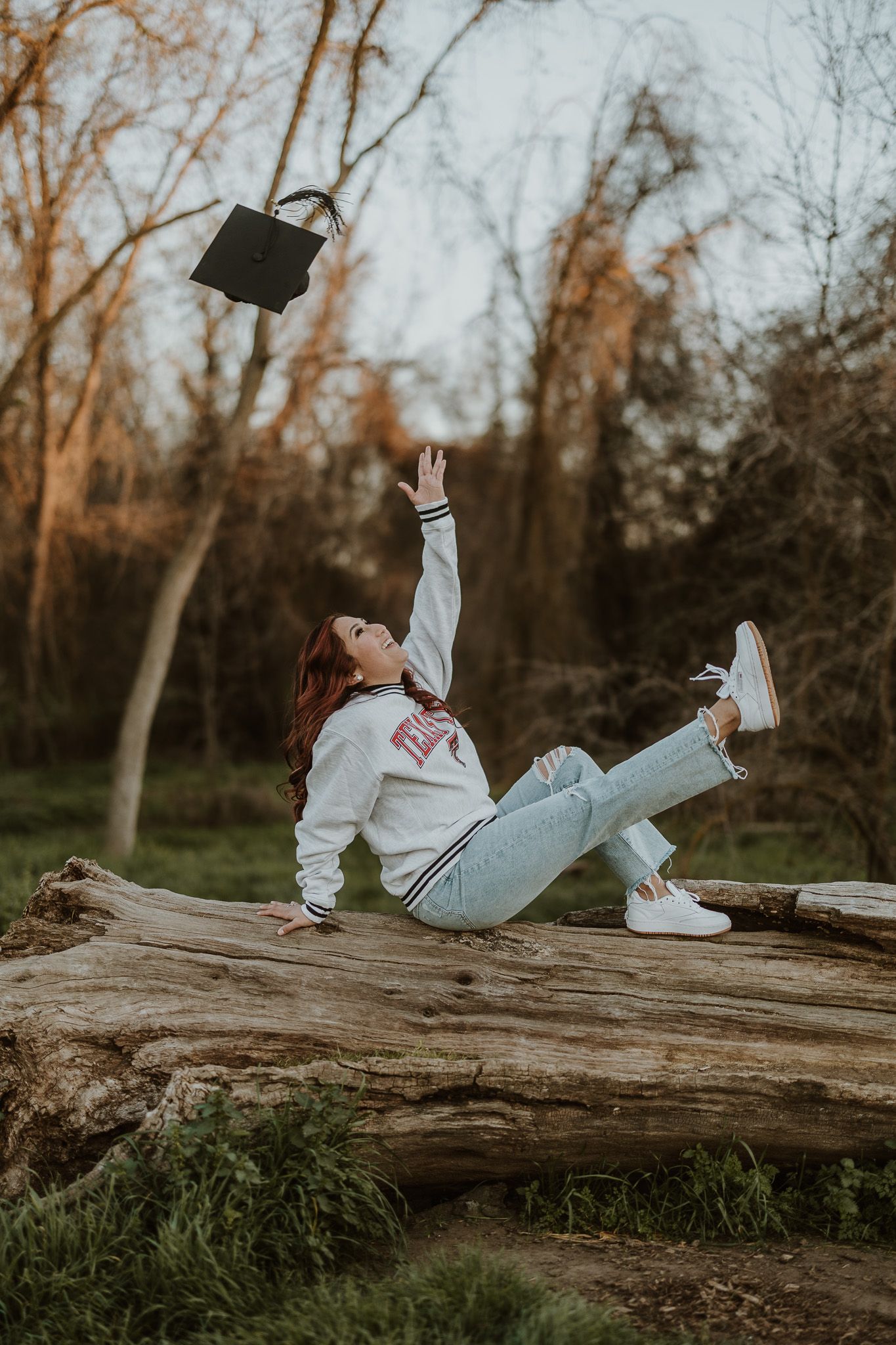 Playful graduation photo of person tossing cap while sitting on fallen log.