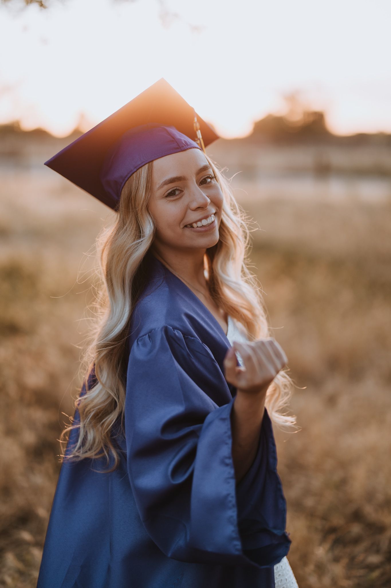Graduate in blue cap and gown smiles radiantly during sunset photo session.