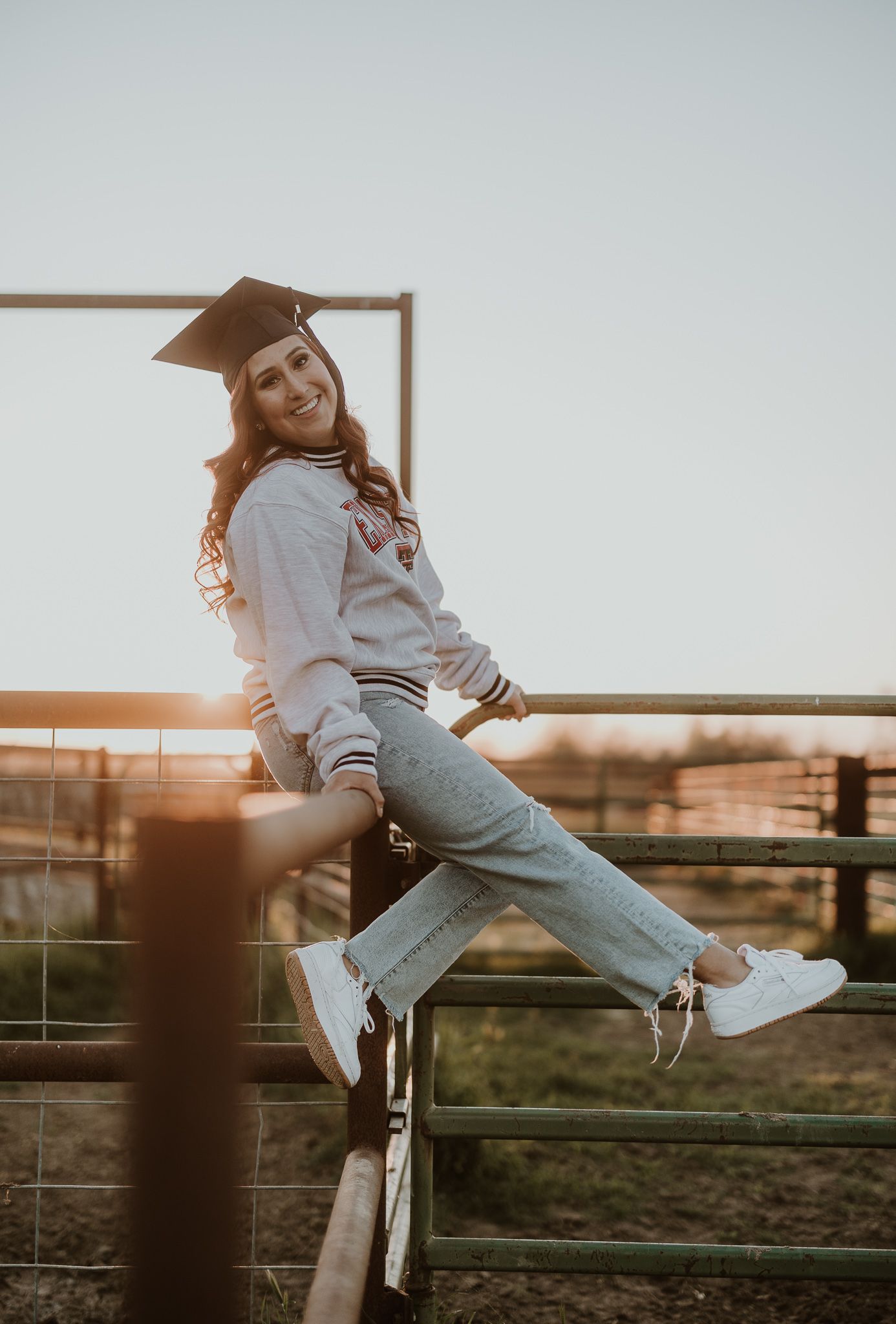 Graduate in casual attire sitting on fence during sunset photo session.