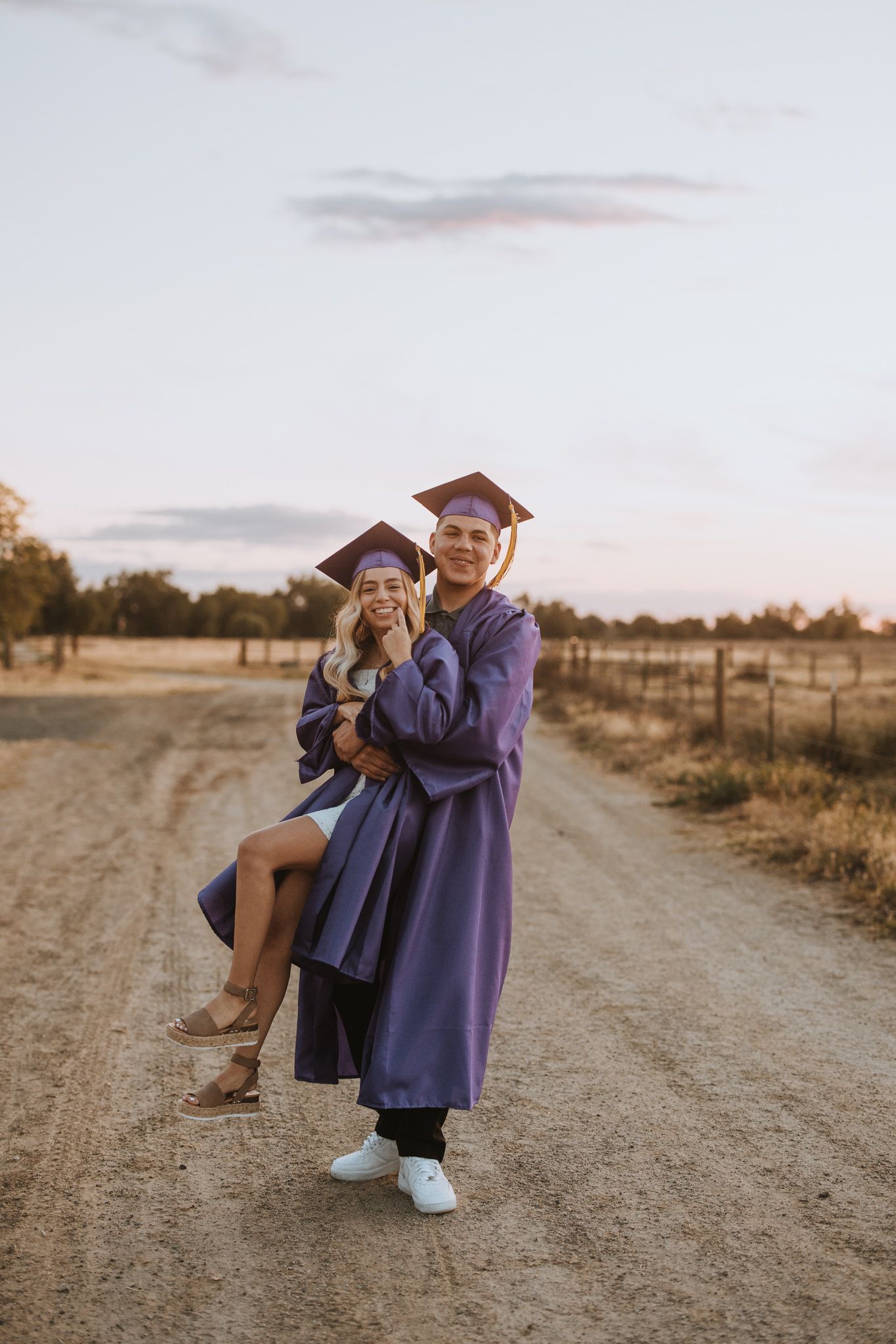 Two graduates in purple caps and gowns share a joyful moment on a dirt path at sunset.