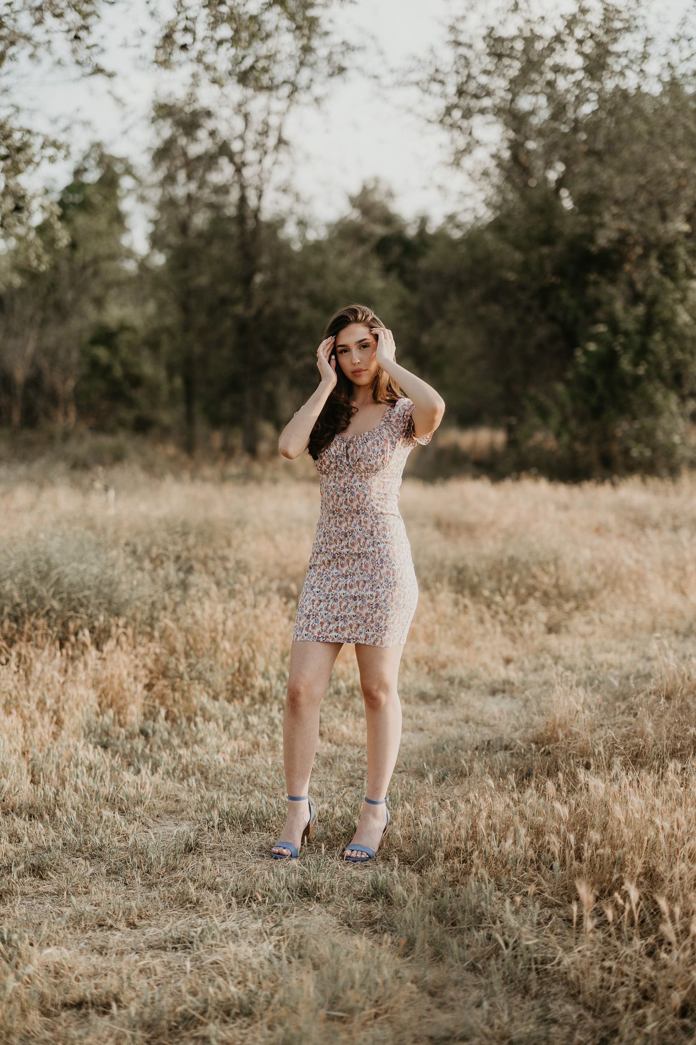 Young woman in fitted floral dress poses in dry grass field during sunset.