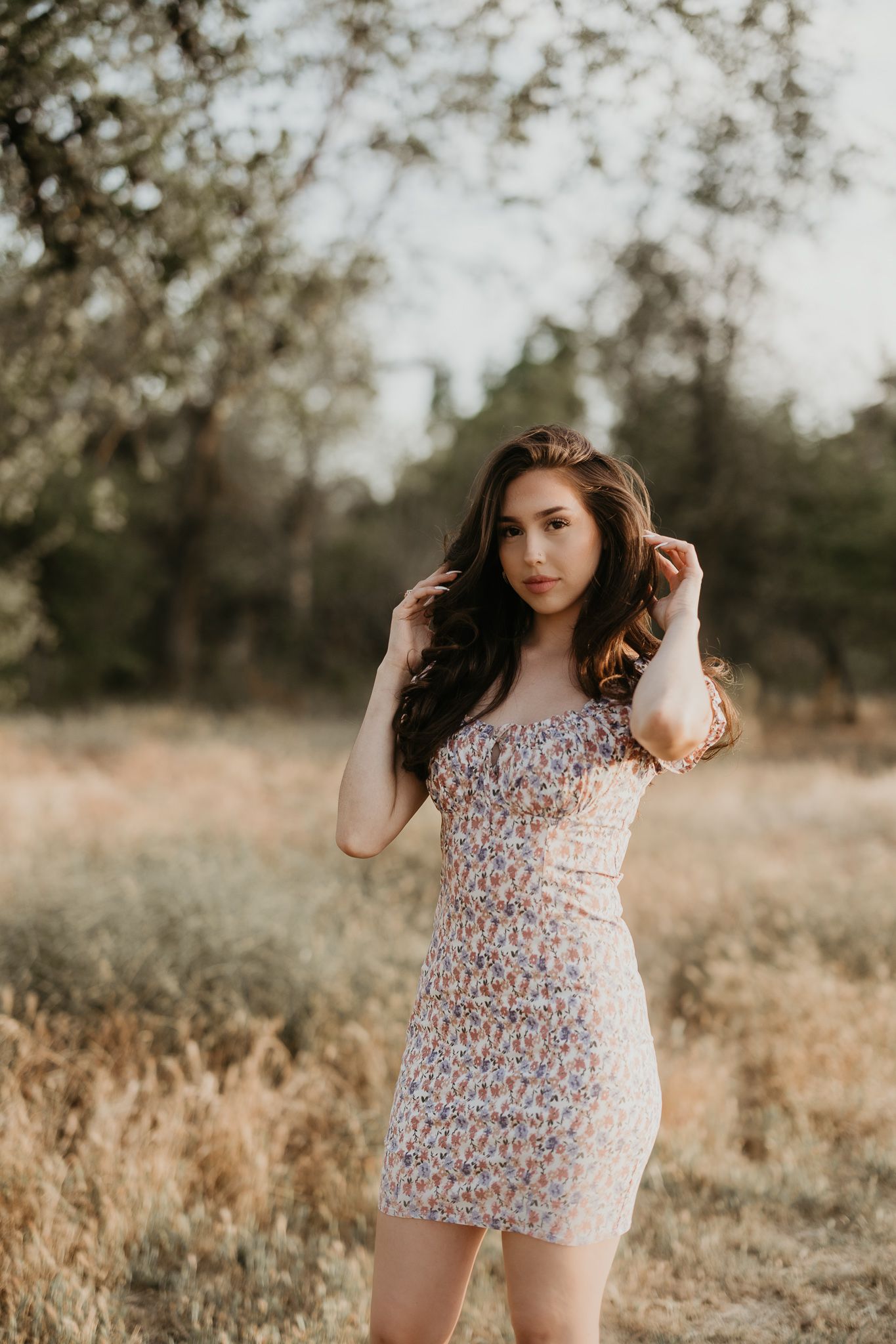 Graduate wearing a floral mini dress poses in a golden field at sunset.