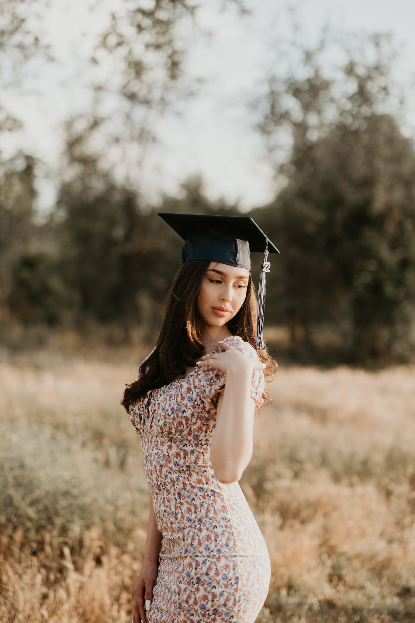 Graduate in floral dress and cap stands in field during golden hour photoshoot.
