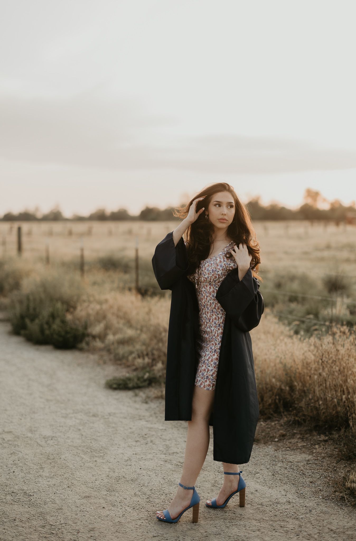 Graduate in fitted dress and graduation gown poses on dirt path at sunset.