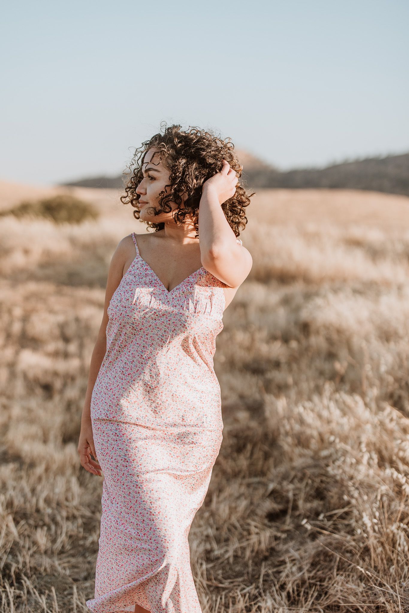 Woman in pink floral dress poses gracefully in field during golden hour.