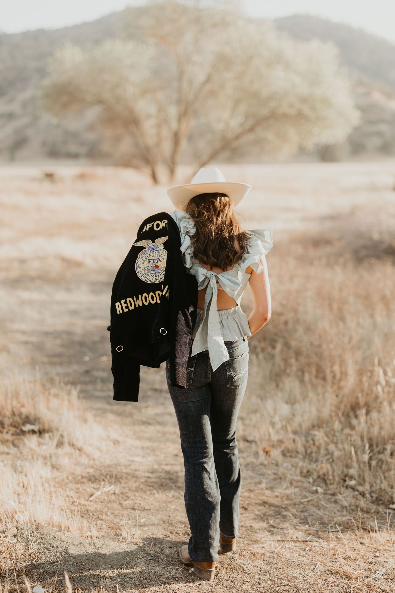 Woman carries decorated denim jacket while walking through field.