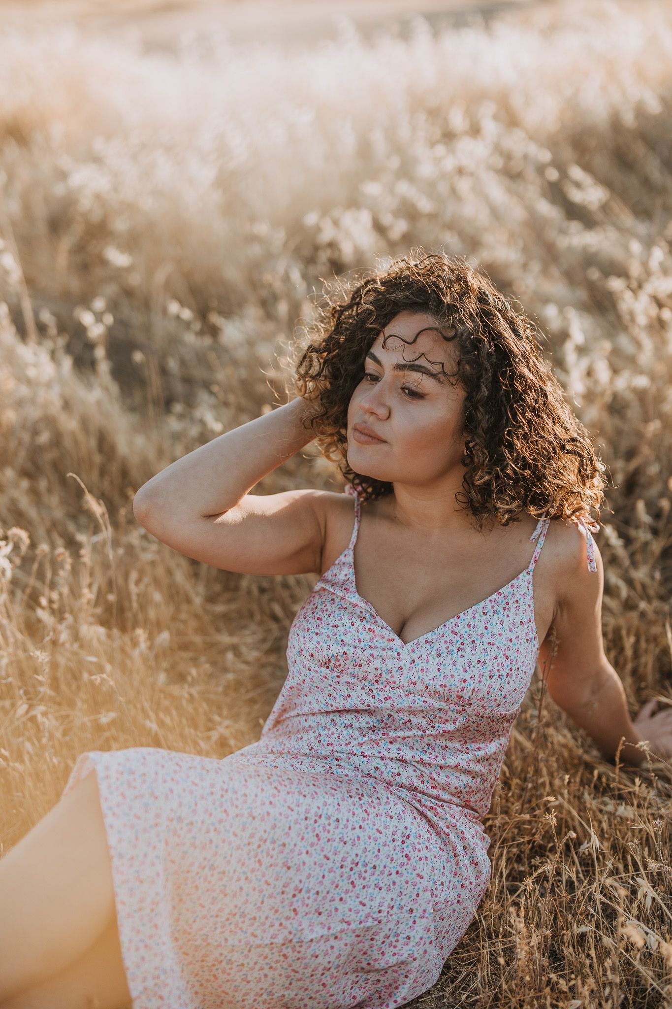 Woman in floral dress reclines in sunlit field during golden hour photoshoot.