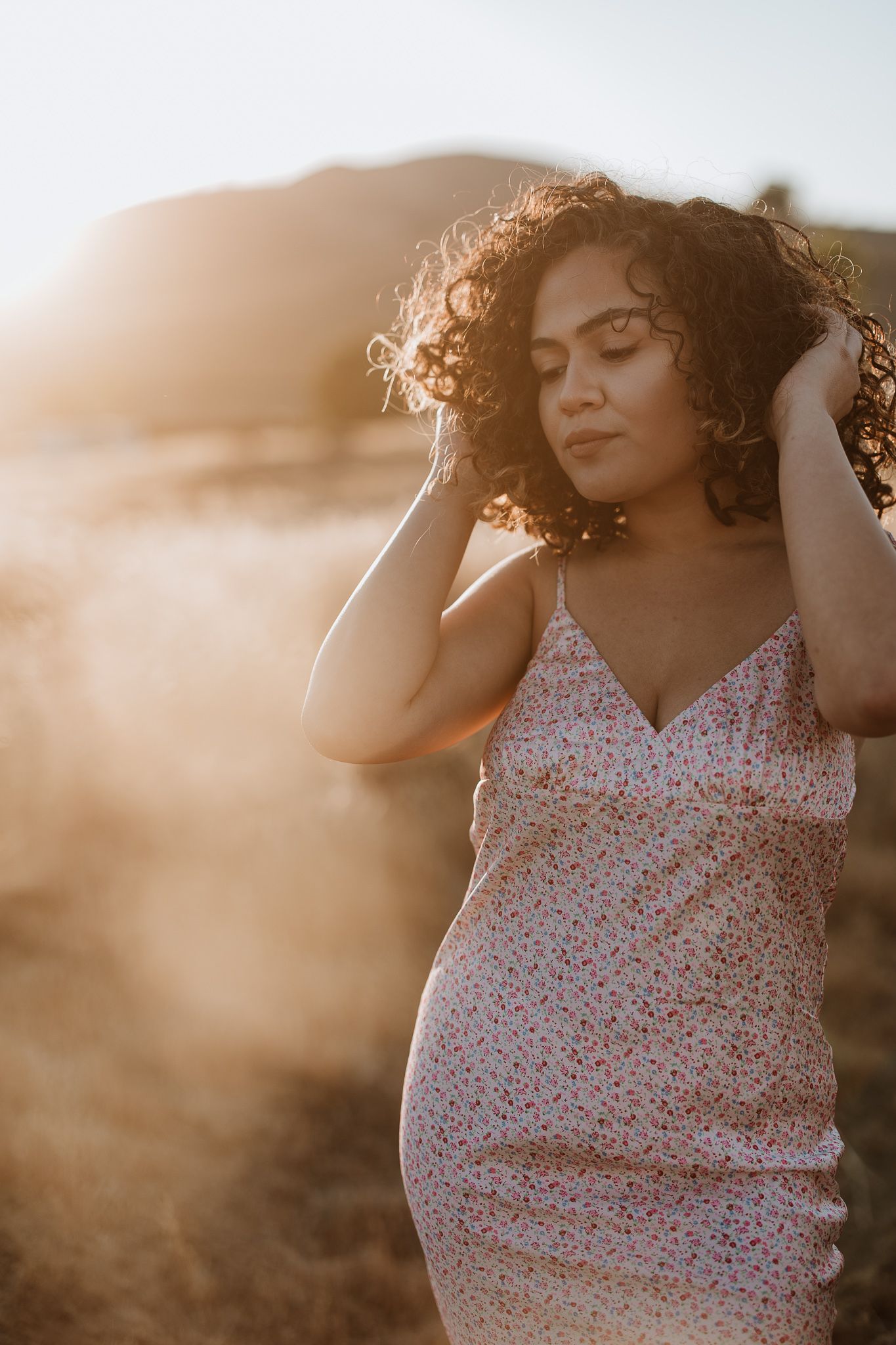 Woman in floral dress stands in golden sunlight with windswept curly hair.