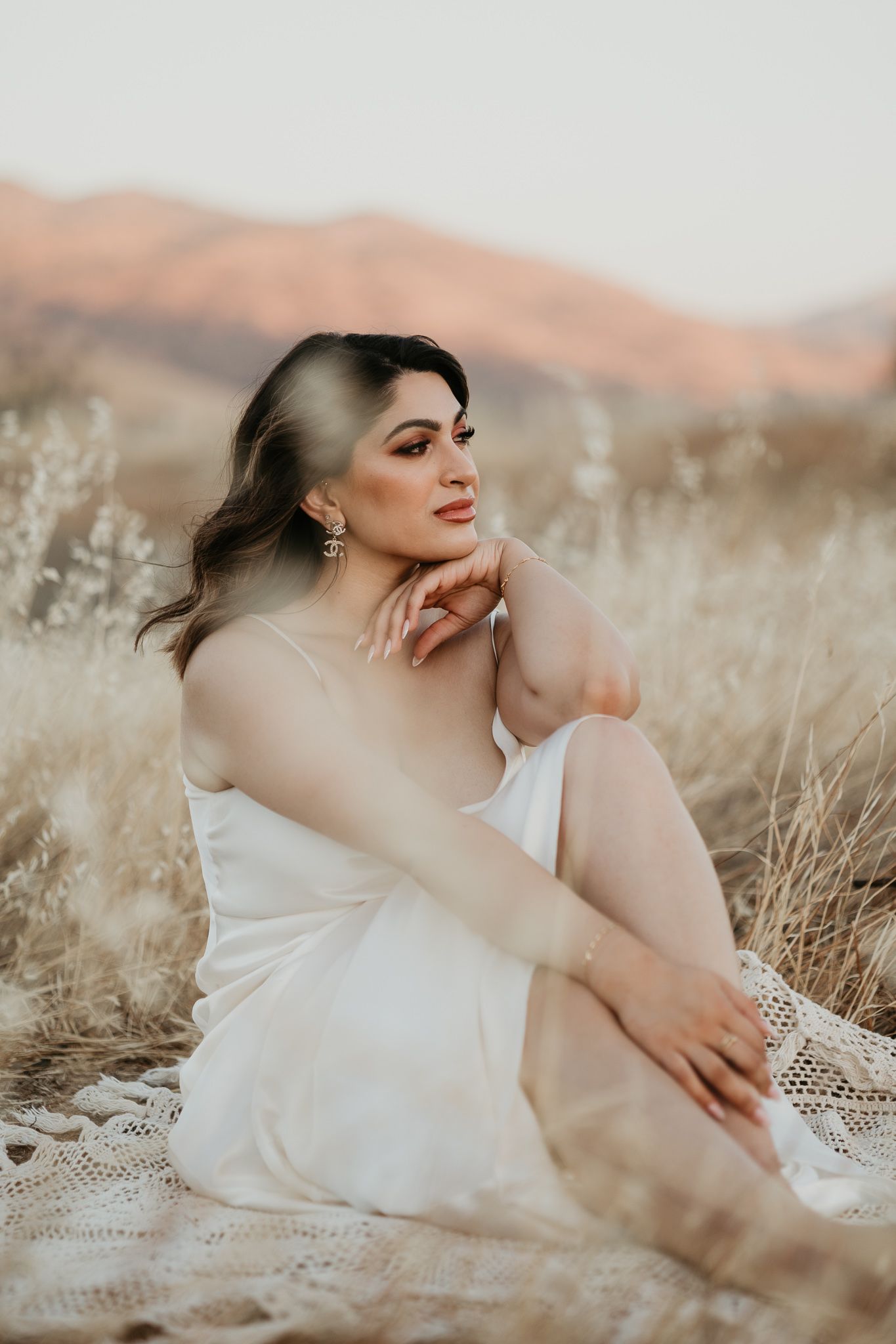 Person in white dress sitting gracefully in tall grass at sunset with mountains in background.