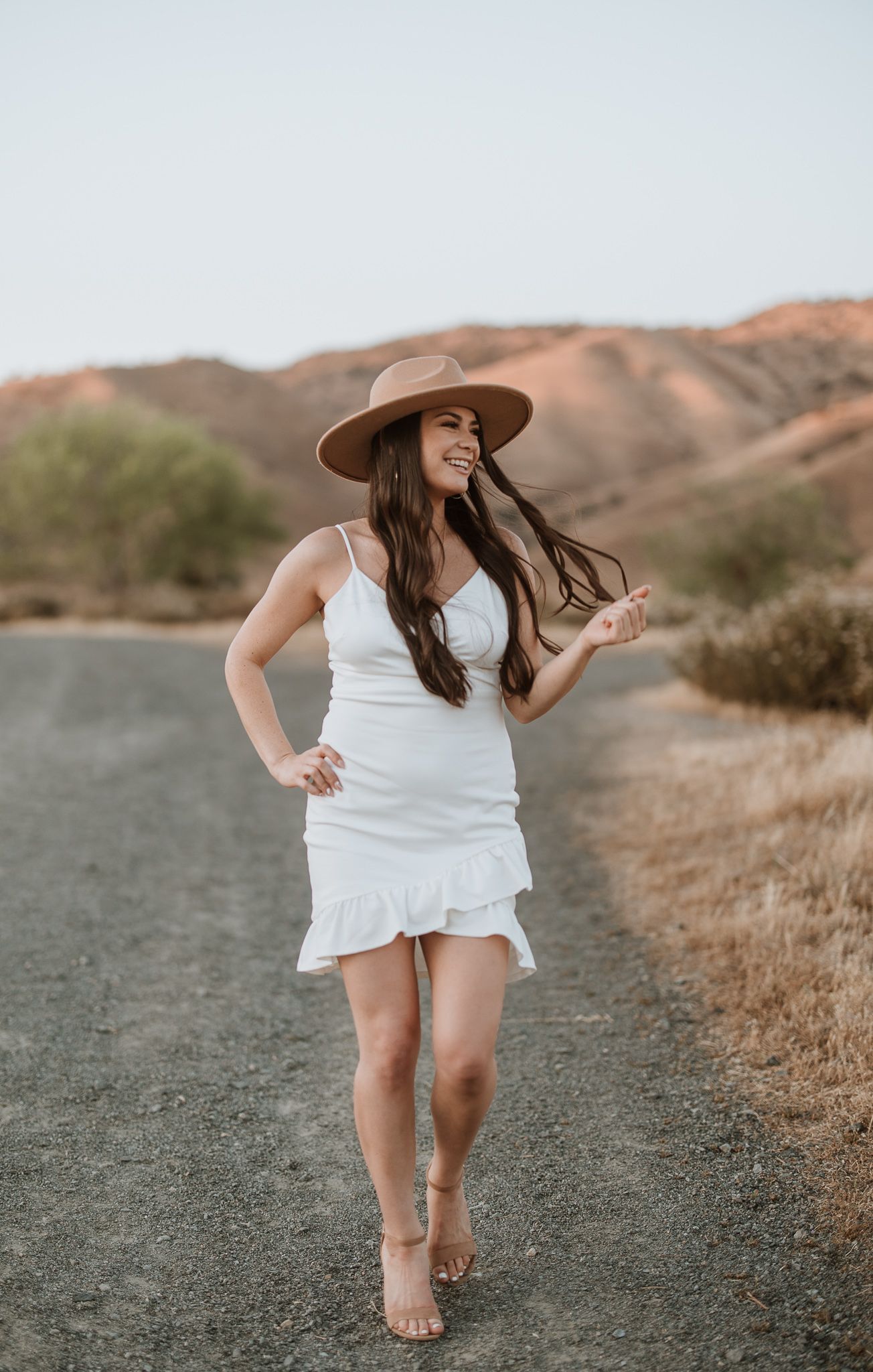 Woman in flowing white dress walking barefoot on desert trail at sunset.