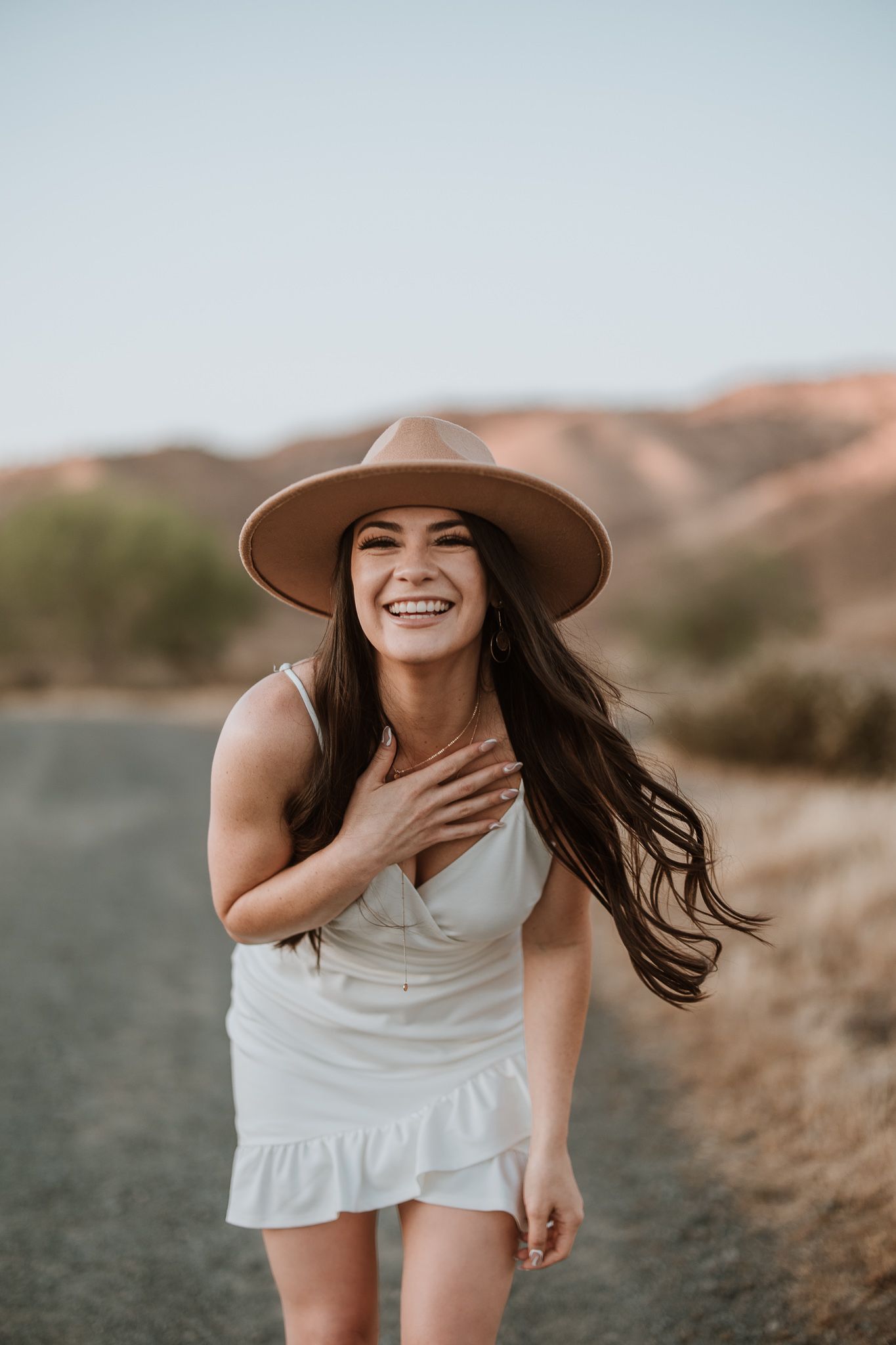 Candid shot of woman in white dress and brown hat laughing on dirt path in desert landscape.