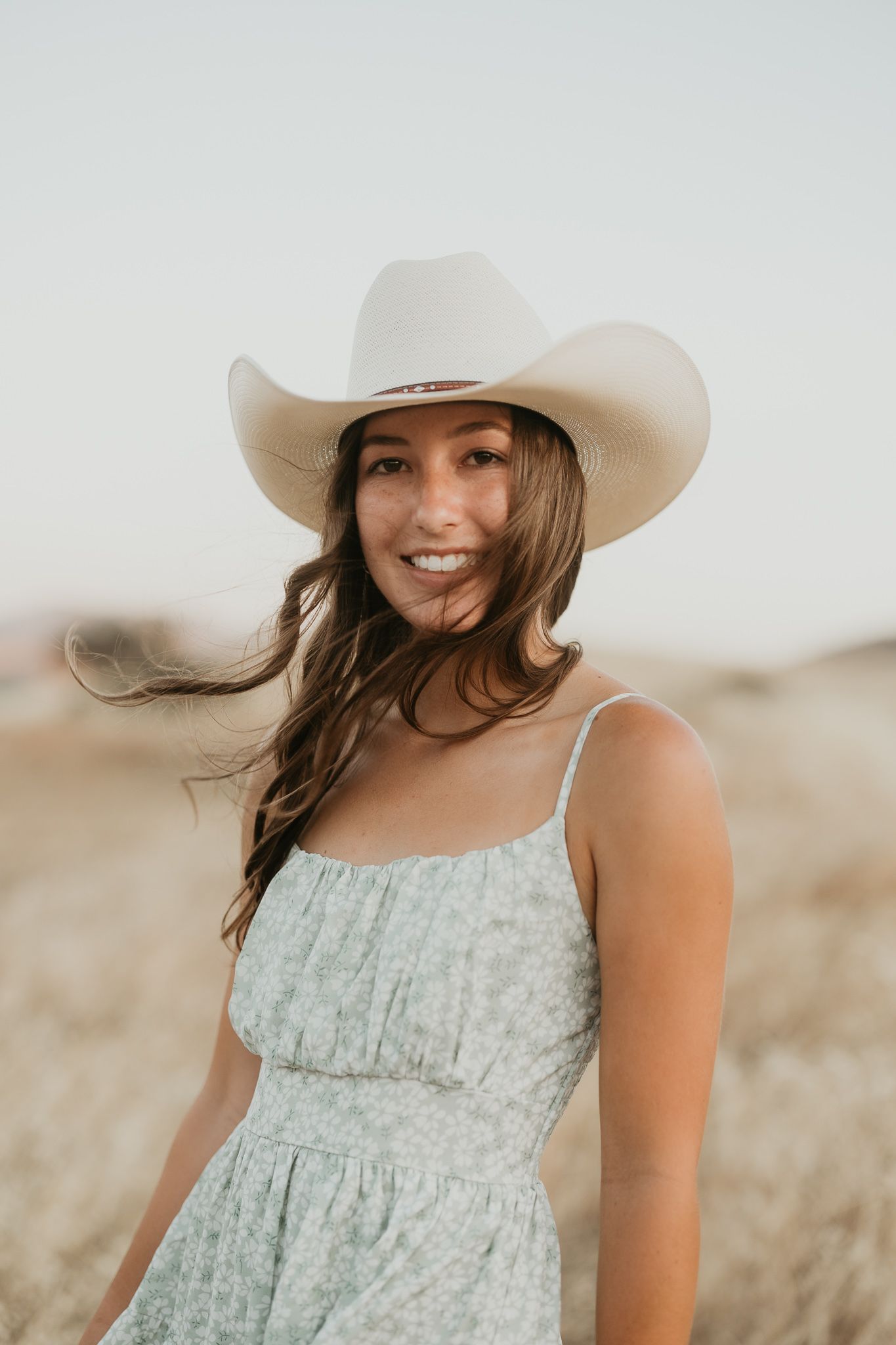 Woman in cowboy hat and sundress smiles at sunset.
