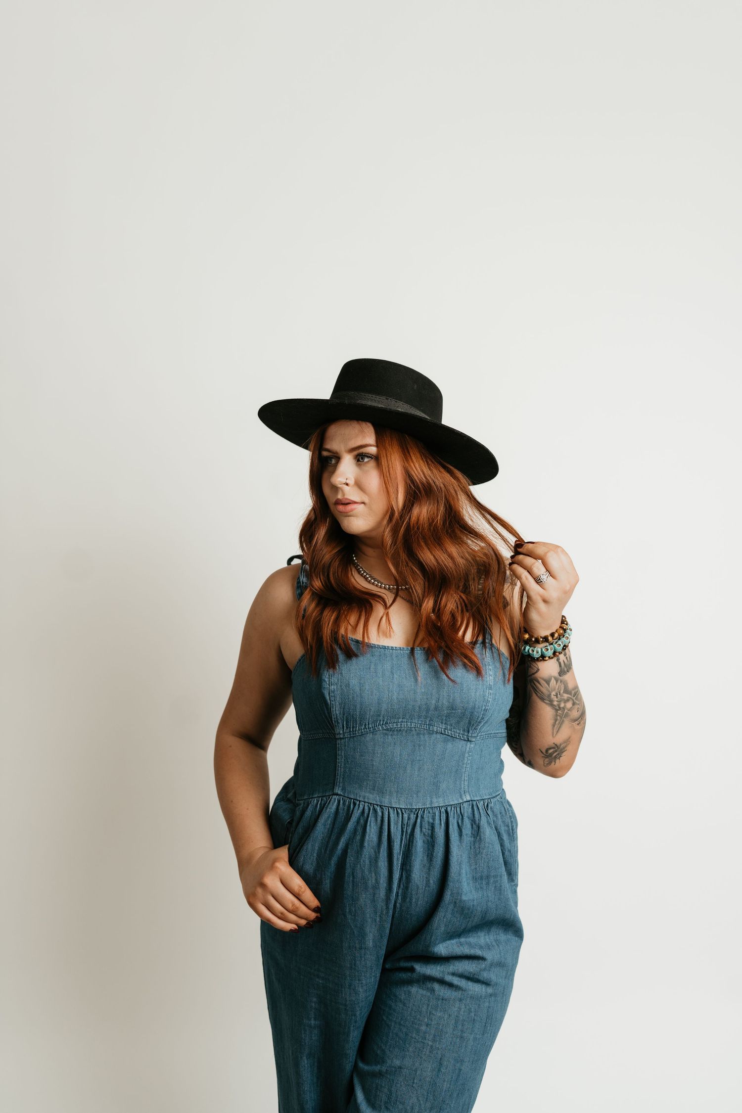 Woman in denim jumpsuit and black hat poses against white background.
