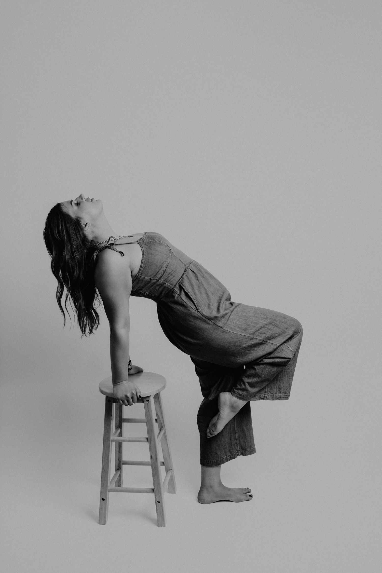 Artistic black and white photo of woman in jumpsuit leaning back on wooden stool.