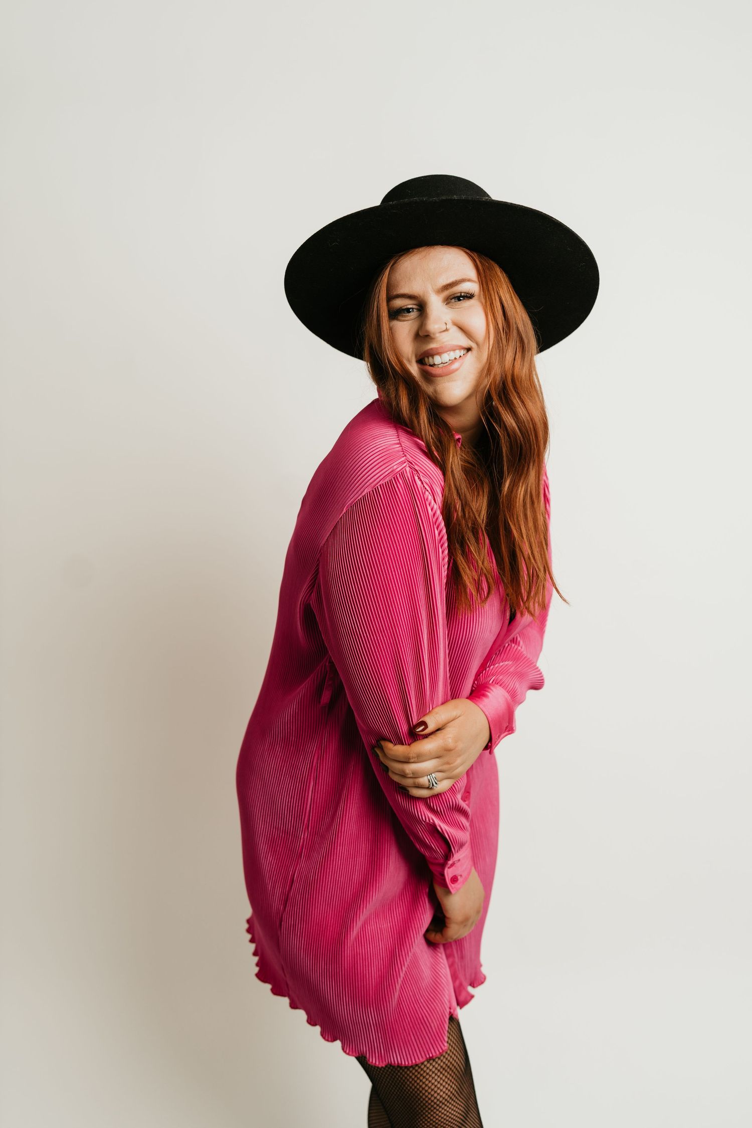 Woman in pink dress and black hat smiling against white background.