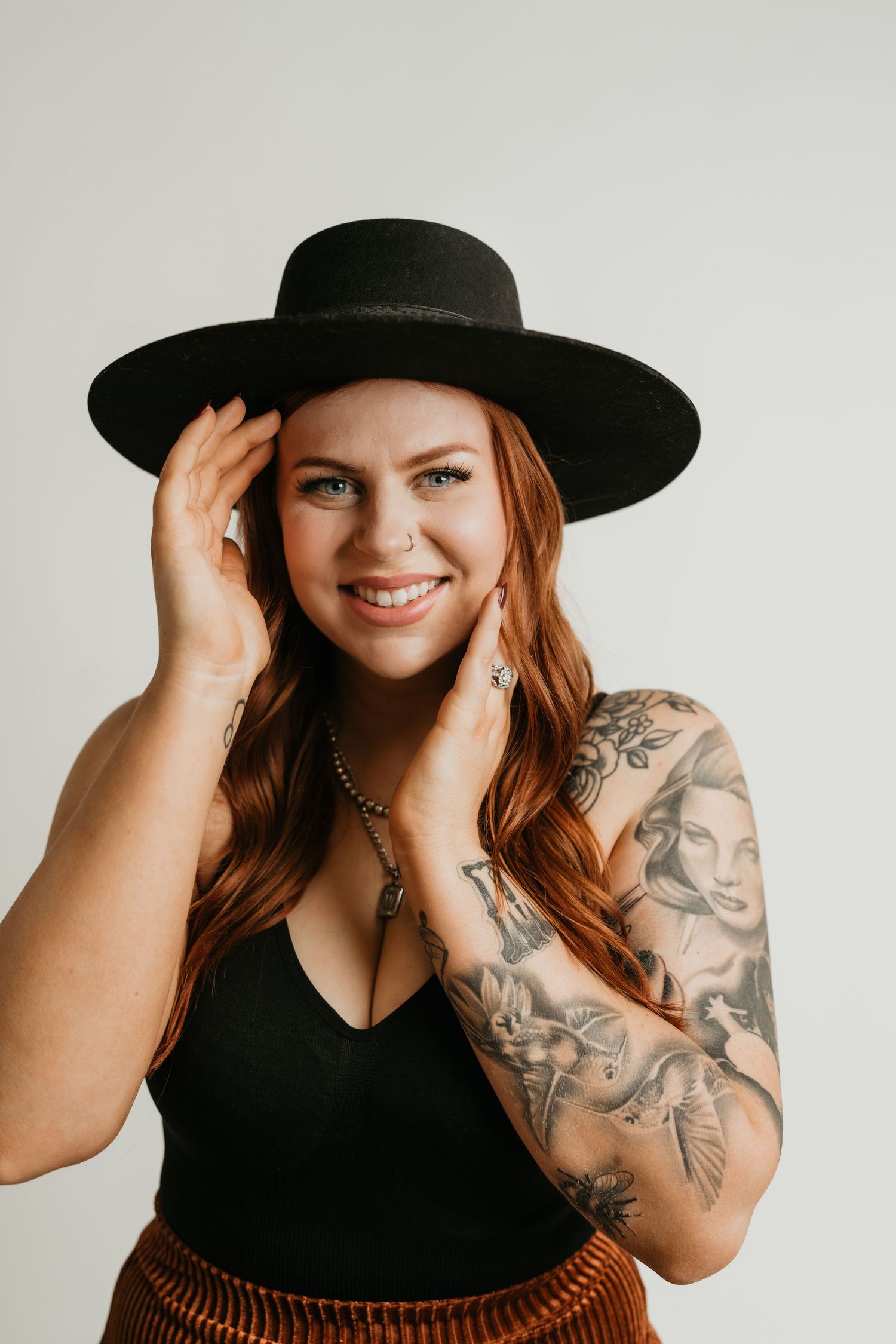 Portrait of a person in a black hat and tank top showing off sleeve tattoos against a white background.