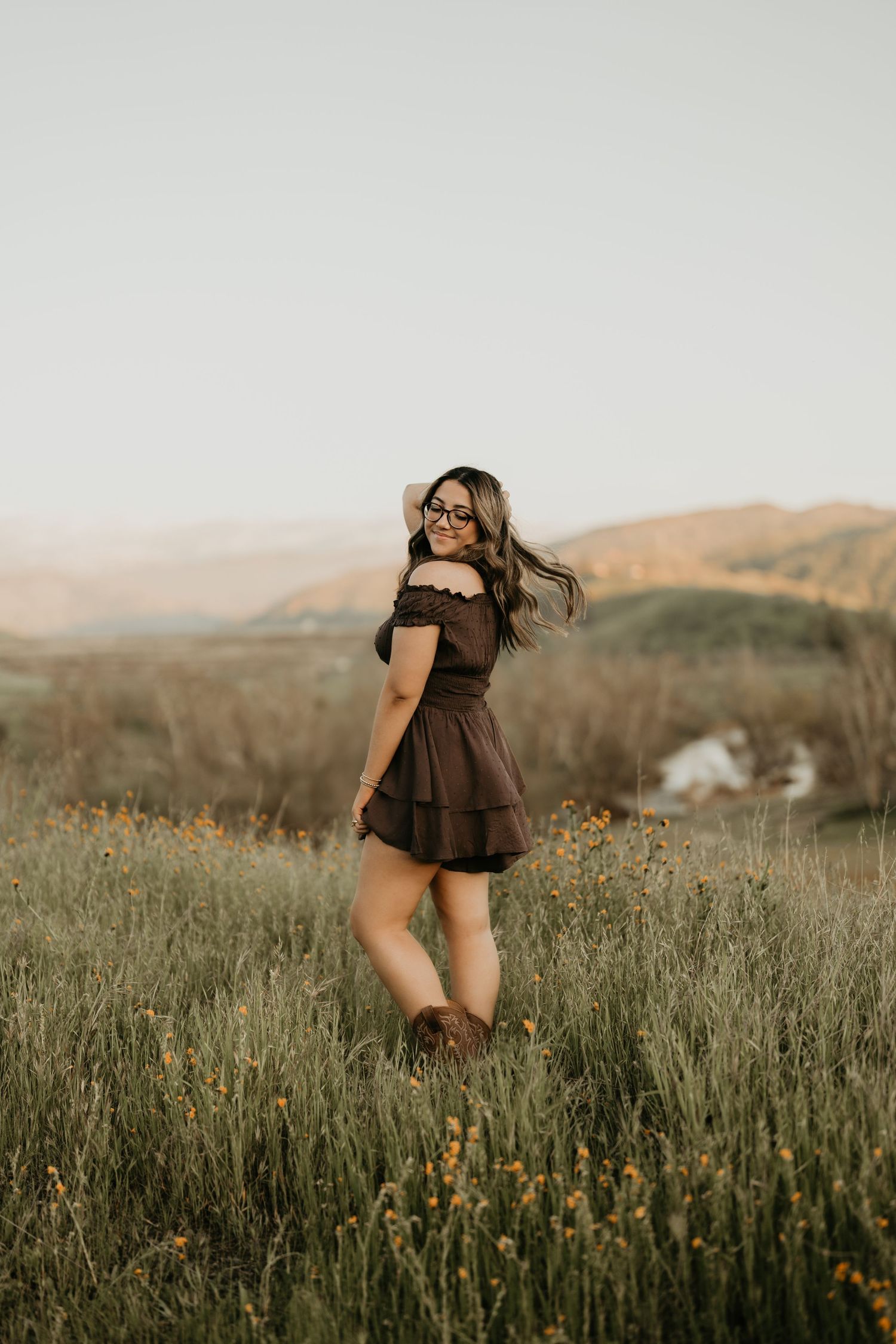 A brown dress twirls against rolling hills and wildflowers in a rustic outdoor setting.