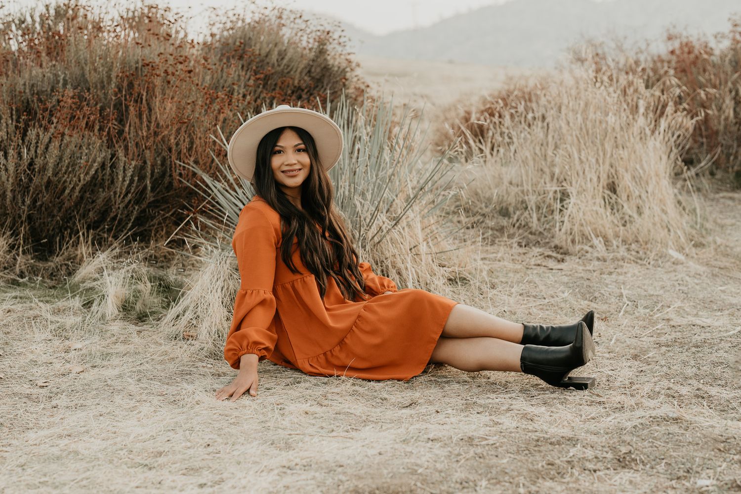 Woman in orange dress and hat poses in dry grass field during golden hour.