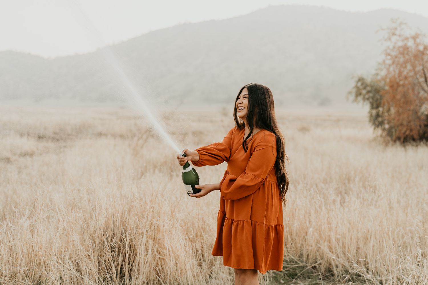 Woman in orange dress spraying champagne in golden field at sunset.