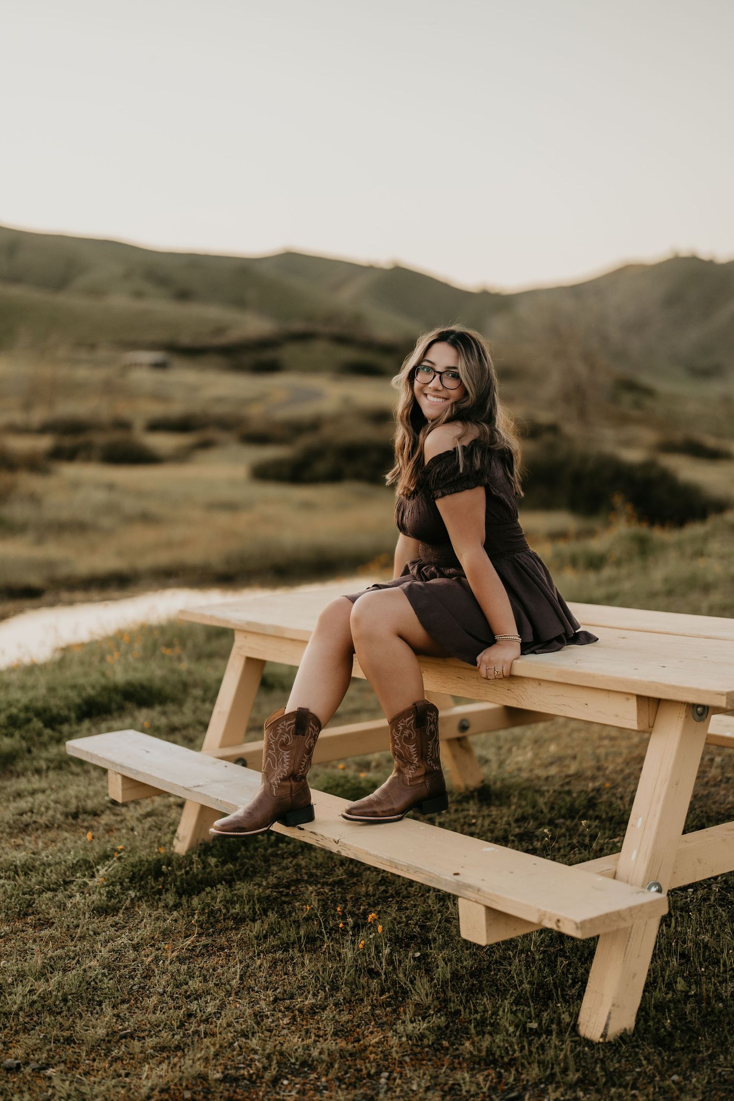 Country-style portrait on wooden picnic table with brown boots and rolling hills backdrop.
