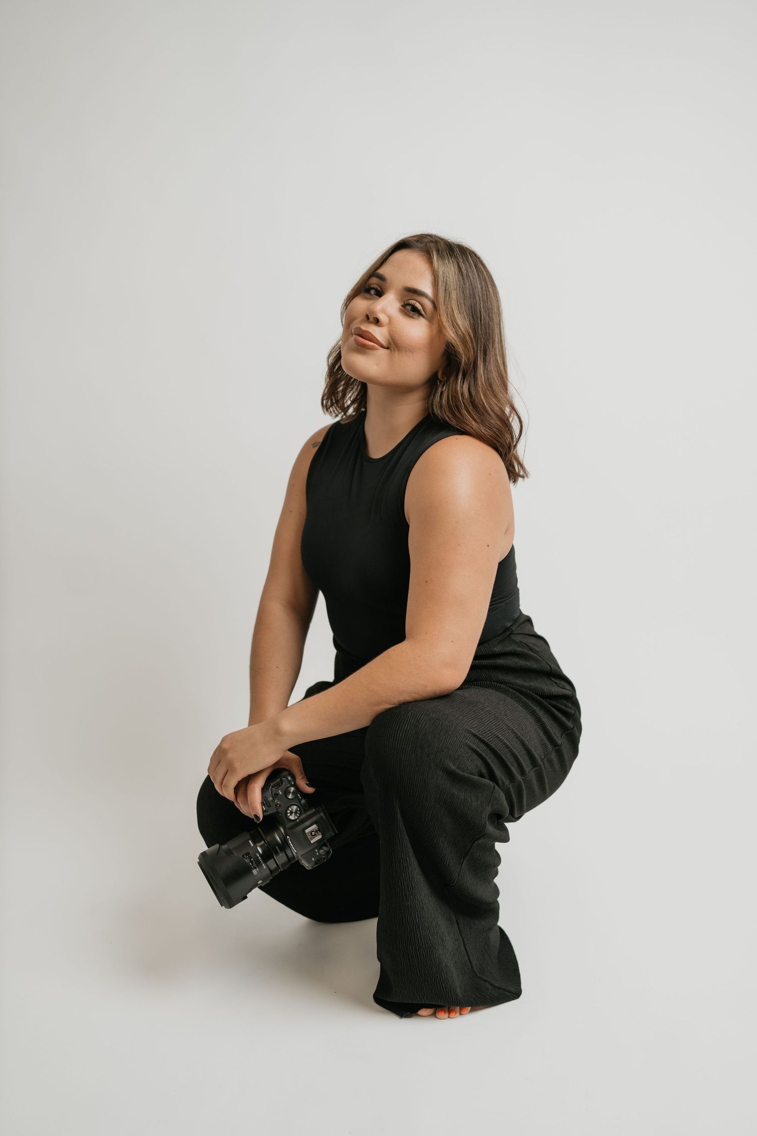 Professional photographer in black outfit crouches with camera against white backdrop.