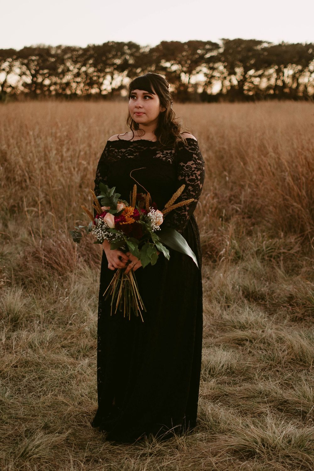 A person in a black lace dress holds a colorful bouquet while standing in a golden field at sunset.