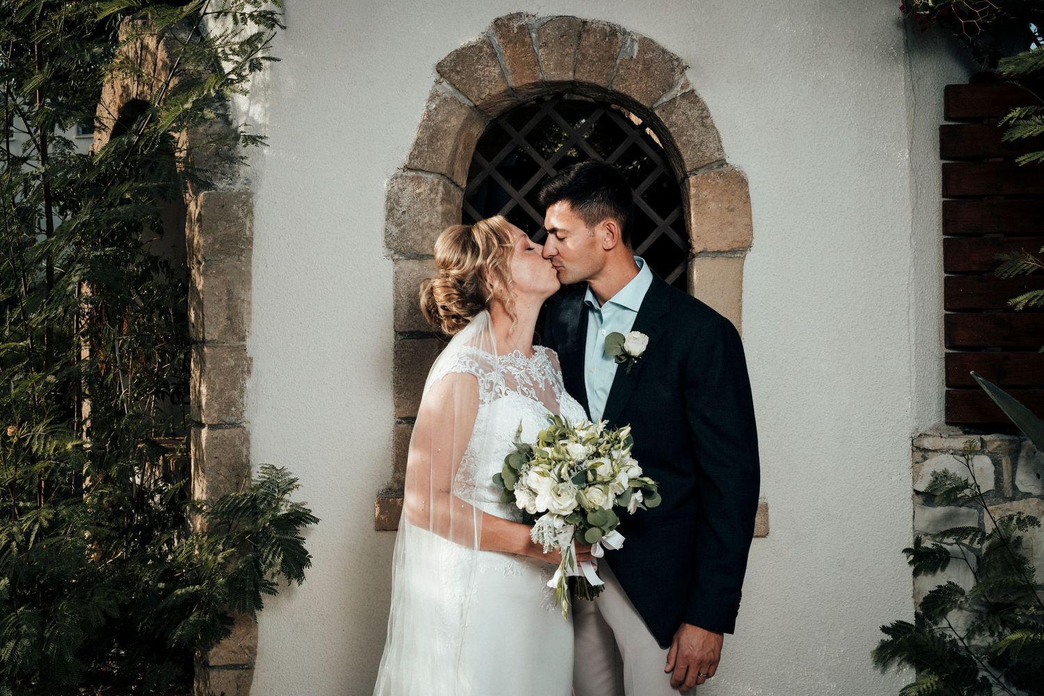 A couple shares a romantic moment under a stone archway with wedding florals.