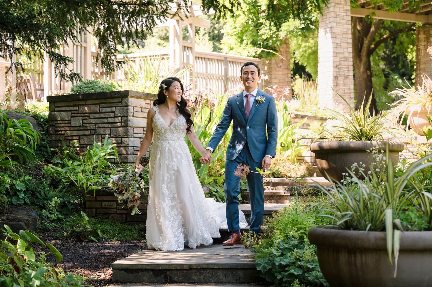 A couple walks hand in hand through a garden setting with stone walls and lush landscaping.