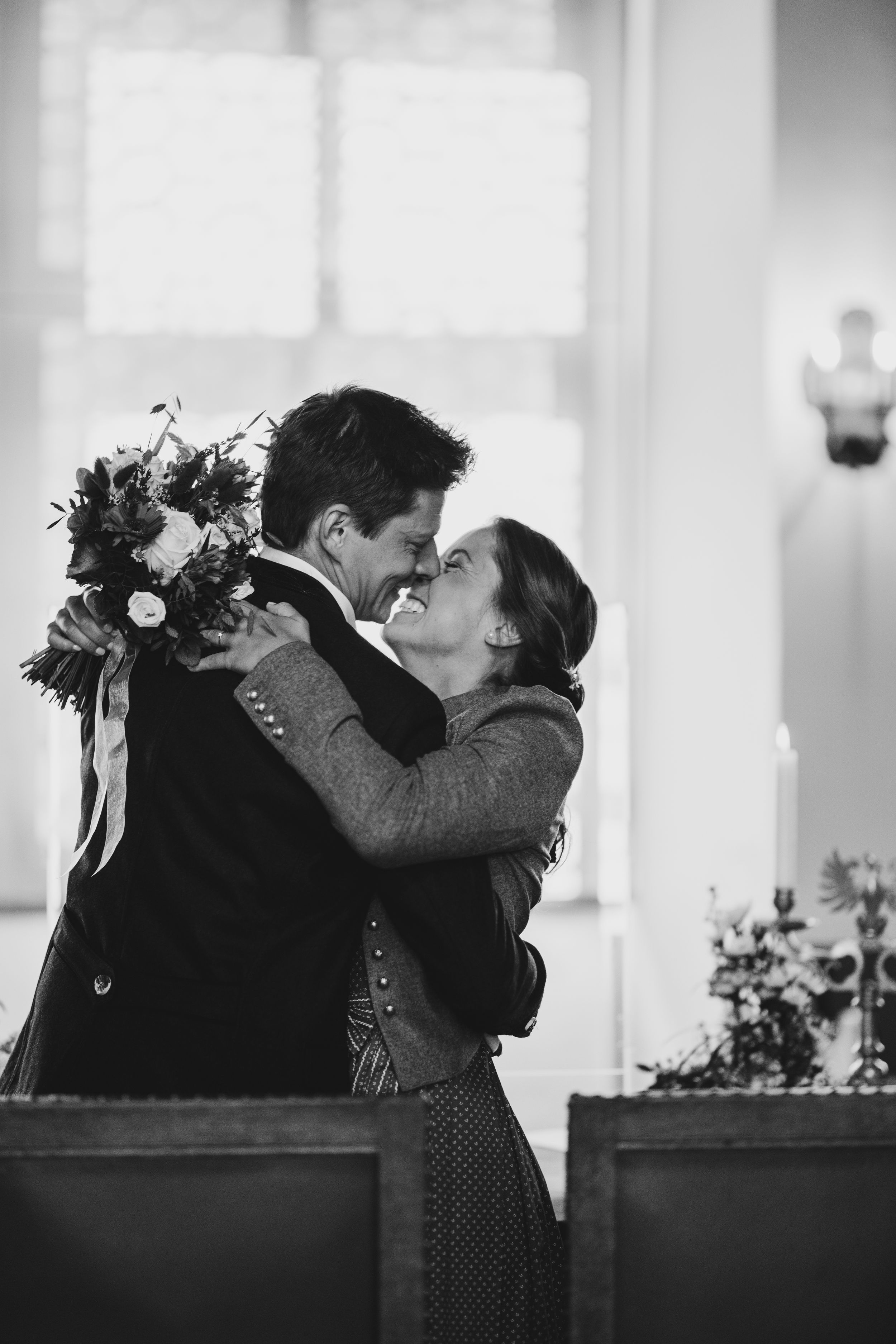 Black and white romantic moment of newlyweds embracing with floral bouquet in church setting.
