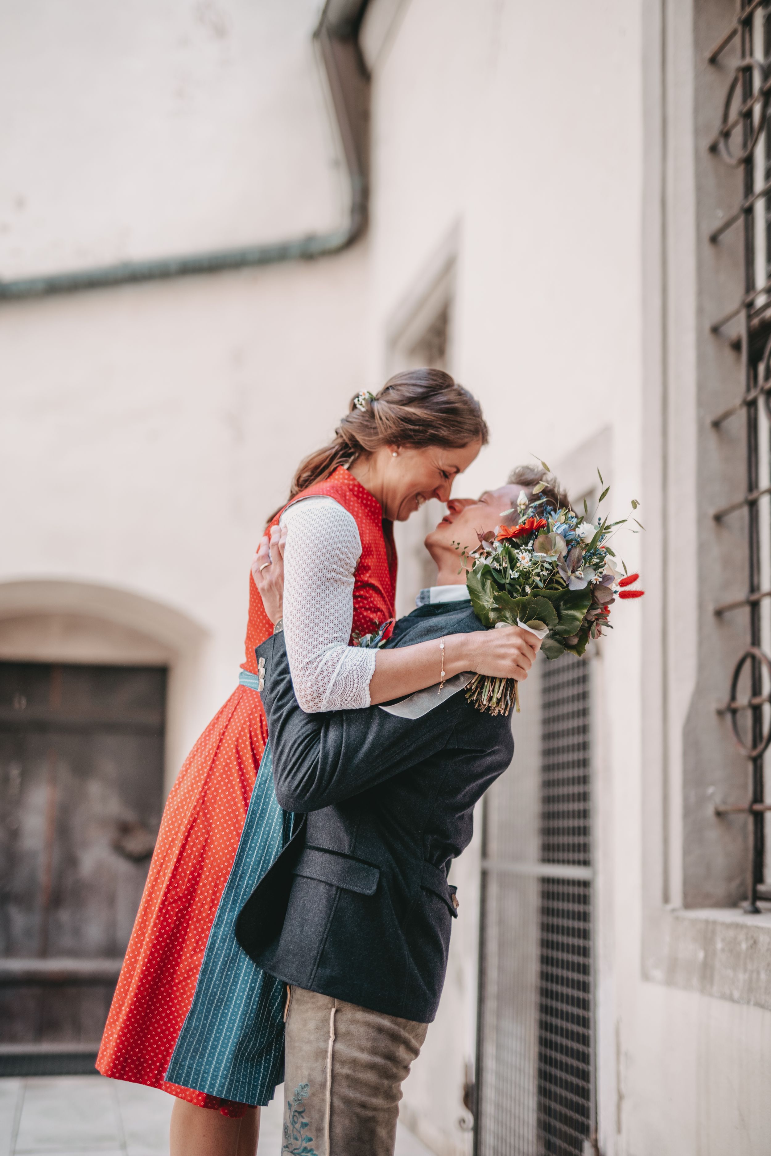 Couple sharing romantic moment with bouquet against white building exterior.