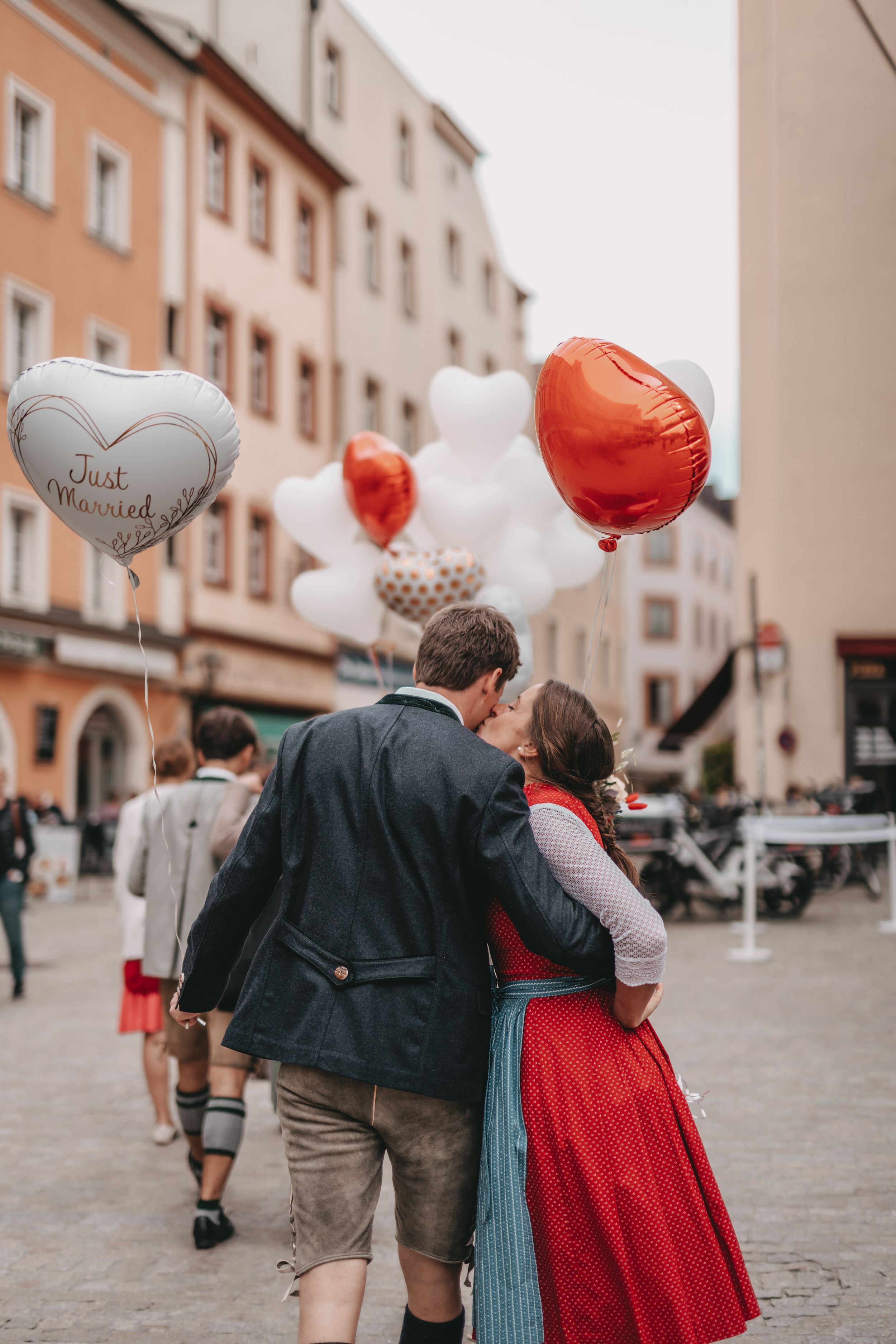 A couple in traditional German dress walking with heart-shaped balloons in a historic town square.