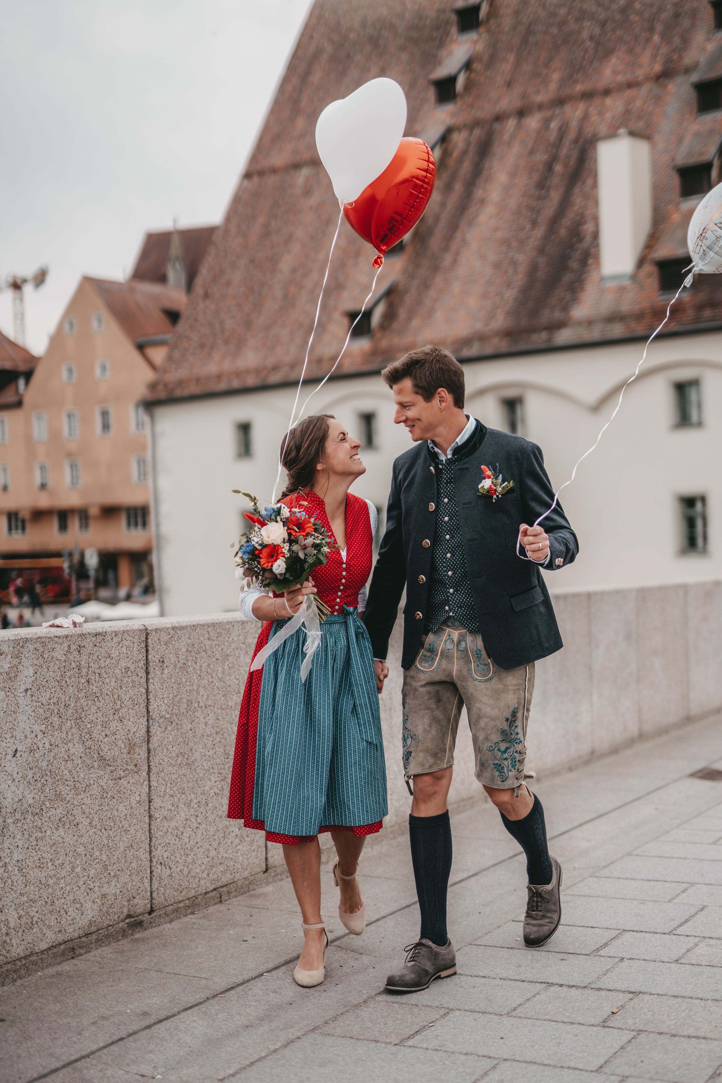 Couple in traditional Bavarian attire walking with balloons against historic architecture.