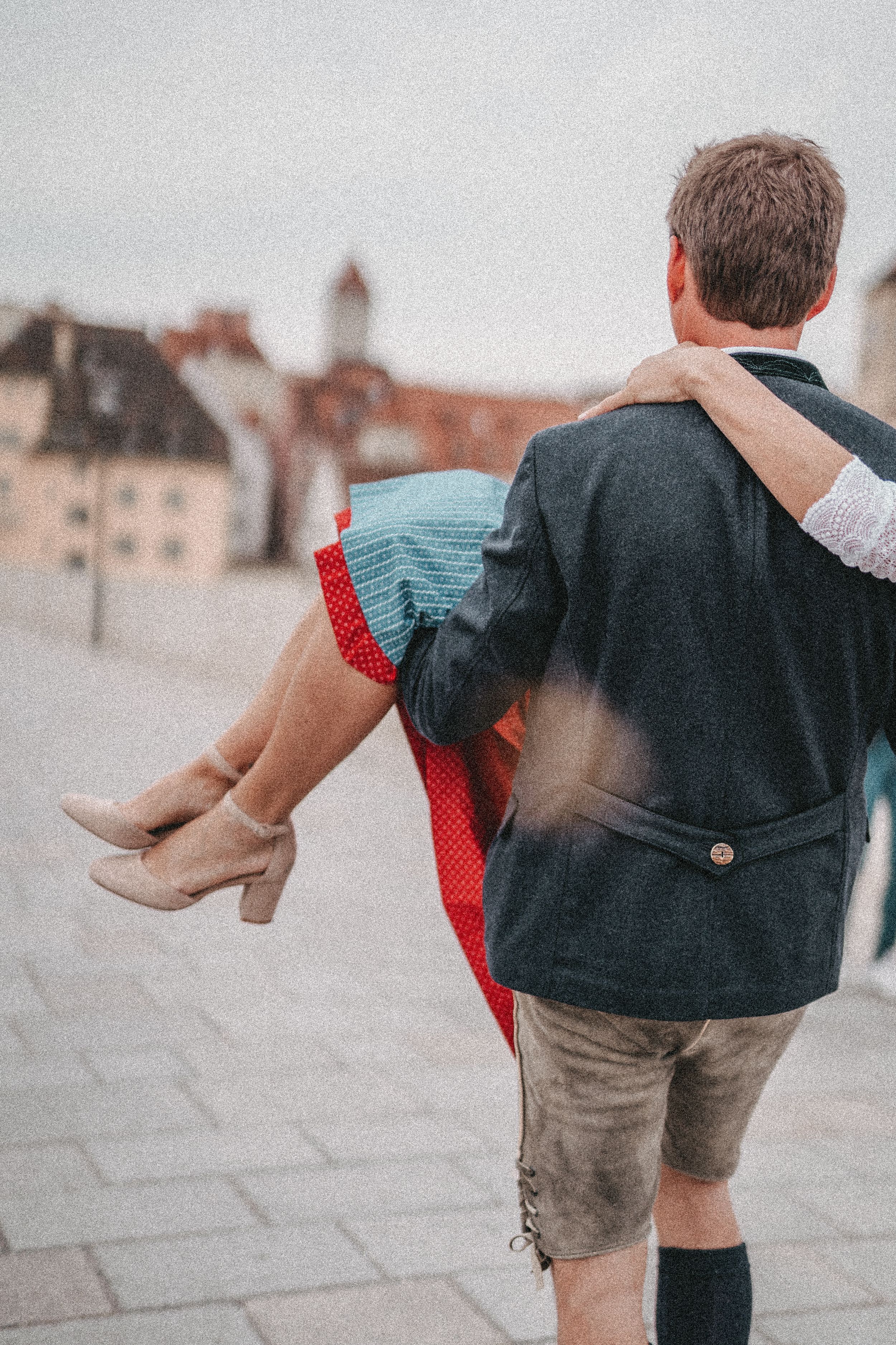 Man in traditional Bavarian clothing carrying woman in dirndl through historic town square.