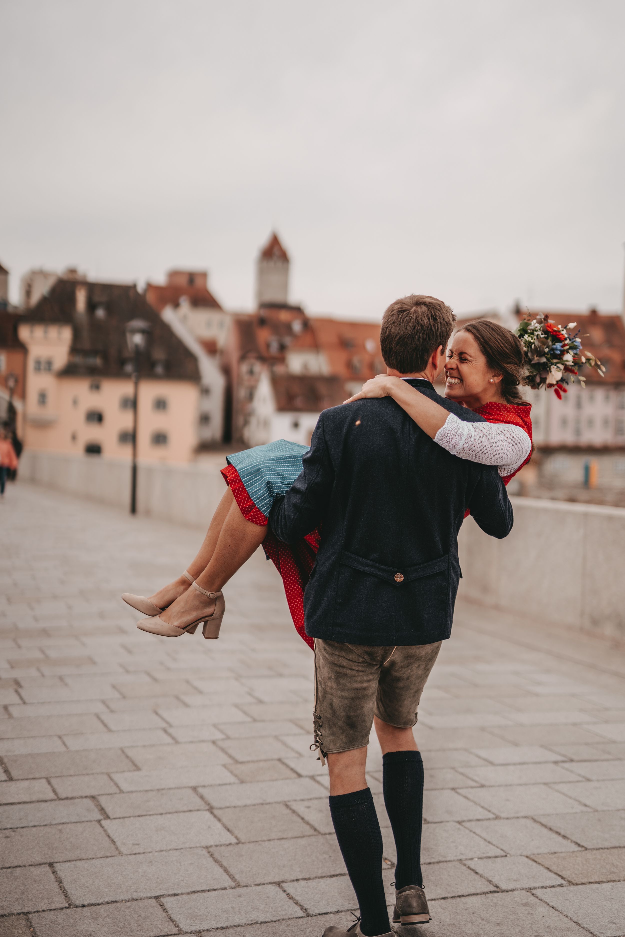 Romantic carry pose in traditional German dress against historic town backdrop.