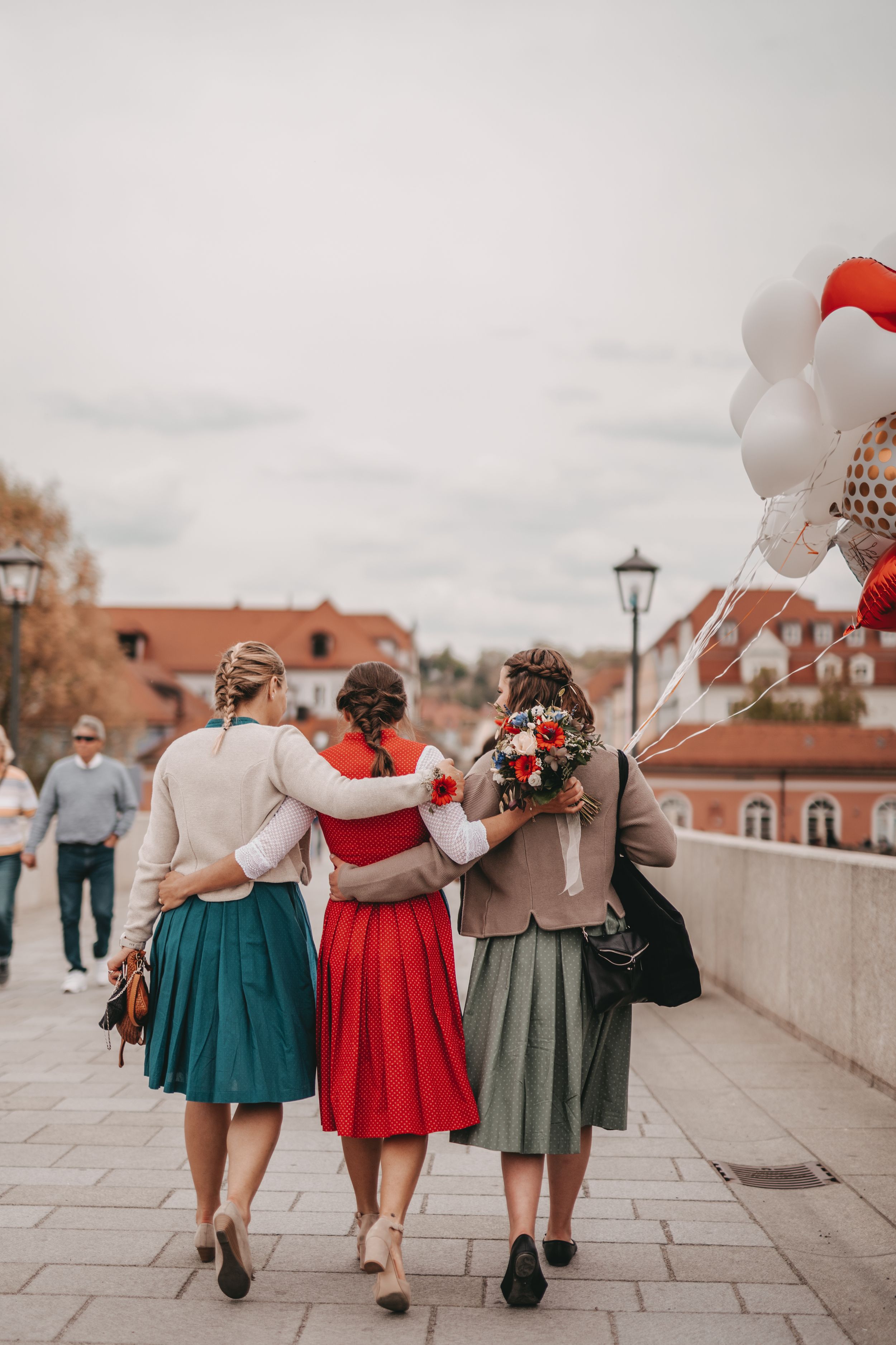 Three friends in vintage-style dresses walk arm-in-arm down a street with red and white balloons in the background.
