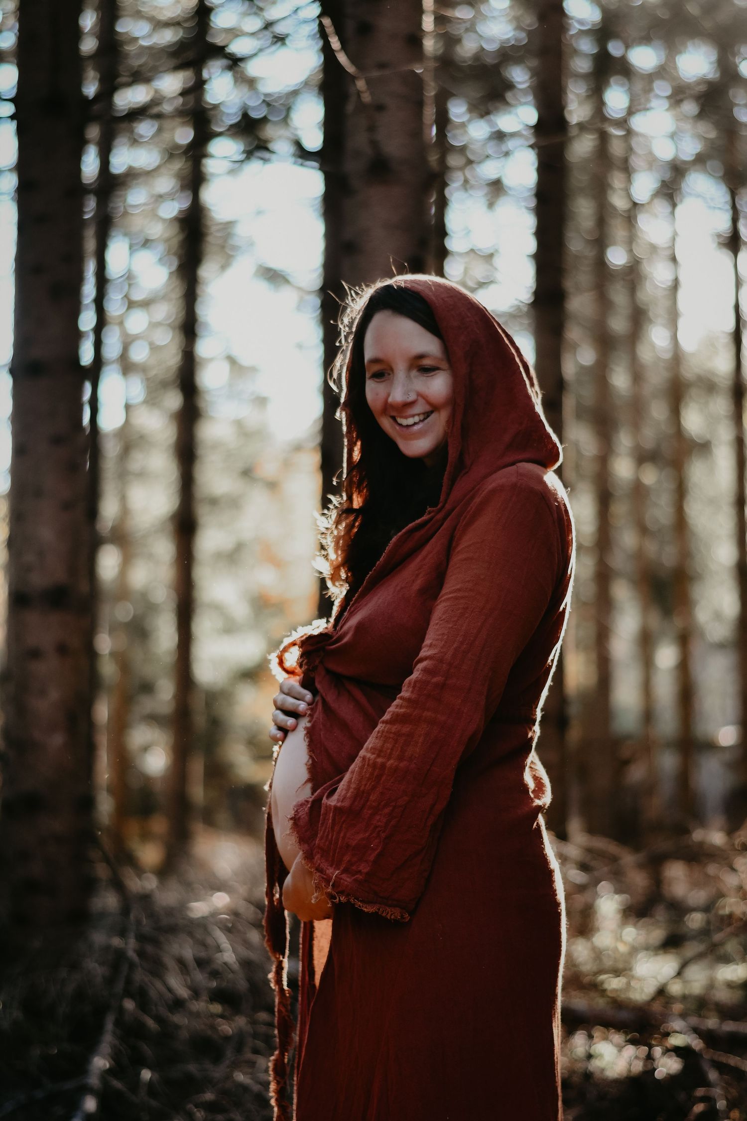 A hooded figure in a burgundy coat stands in a sunlit forest setting.