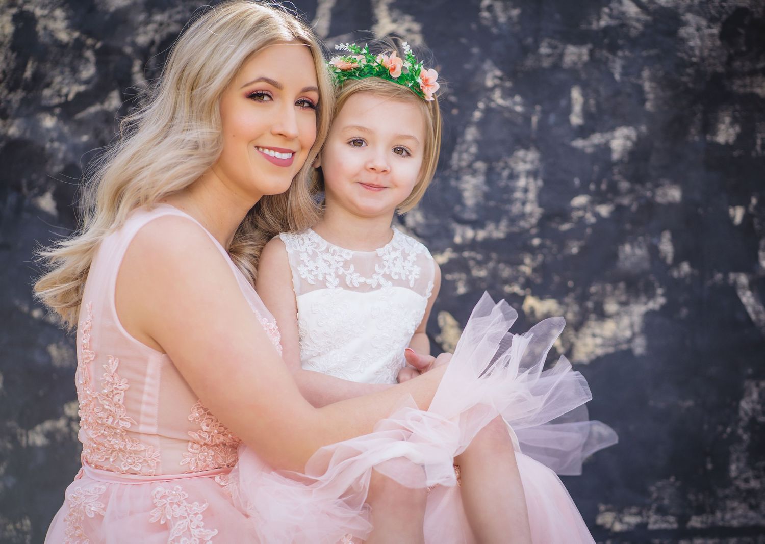A tender portrait of parent and child wearing coordinating pink and white formal dresses against a textured wall.